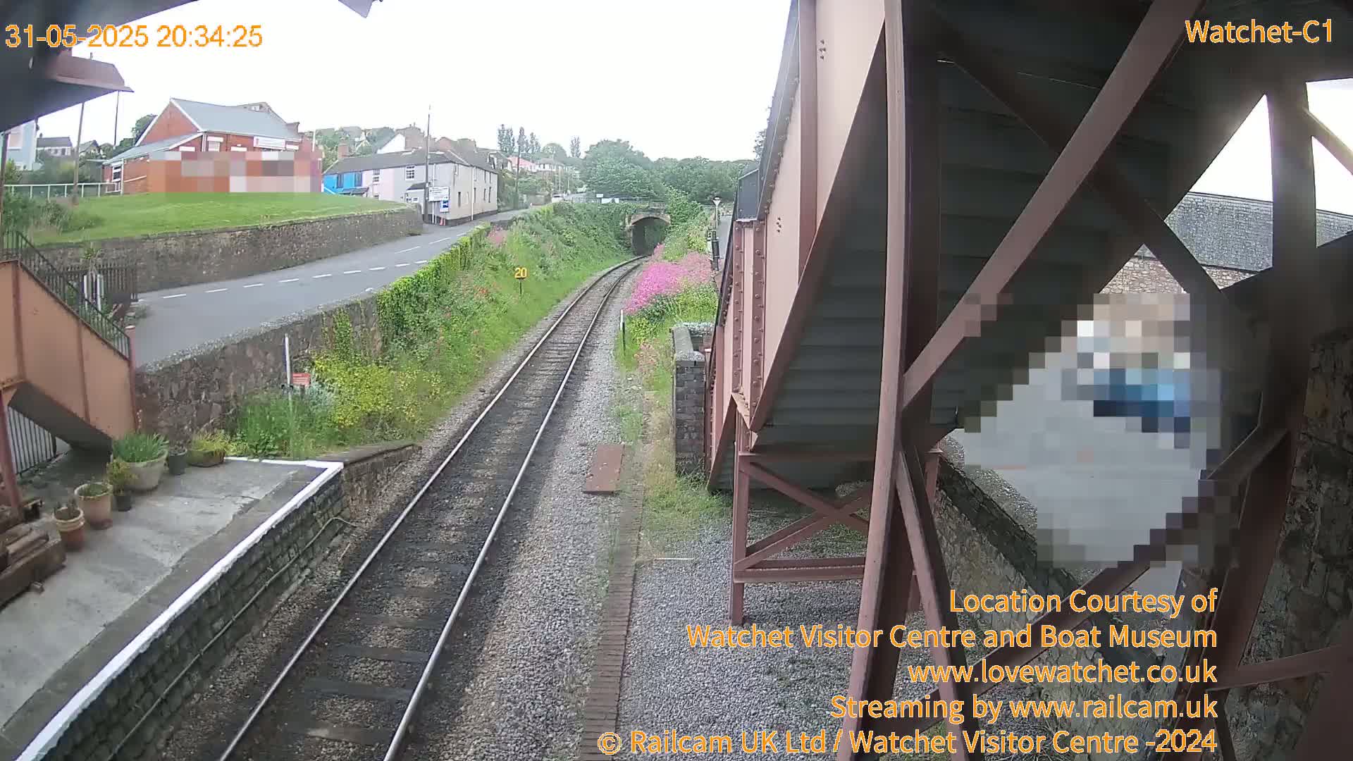 A view from a railway bridge shows train tracks leading to a tunnel, with a small town and overcast sky in the background.