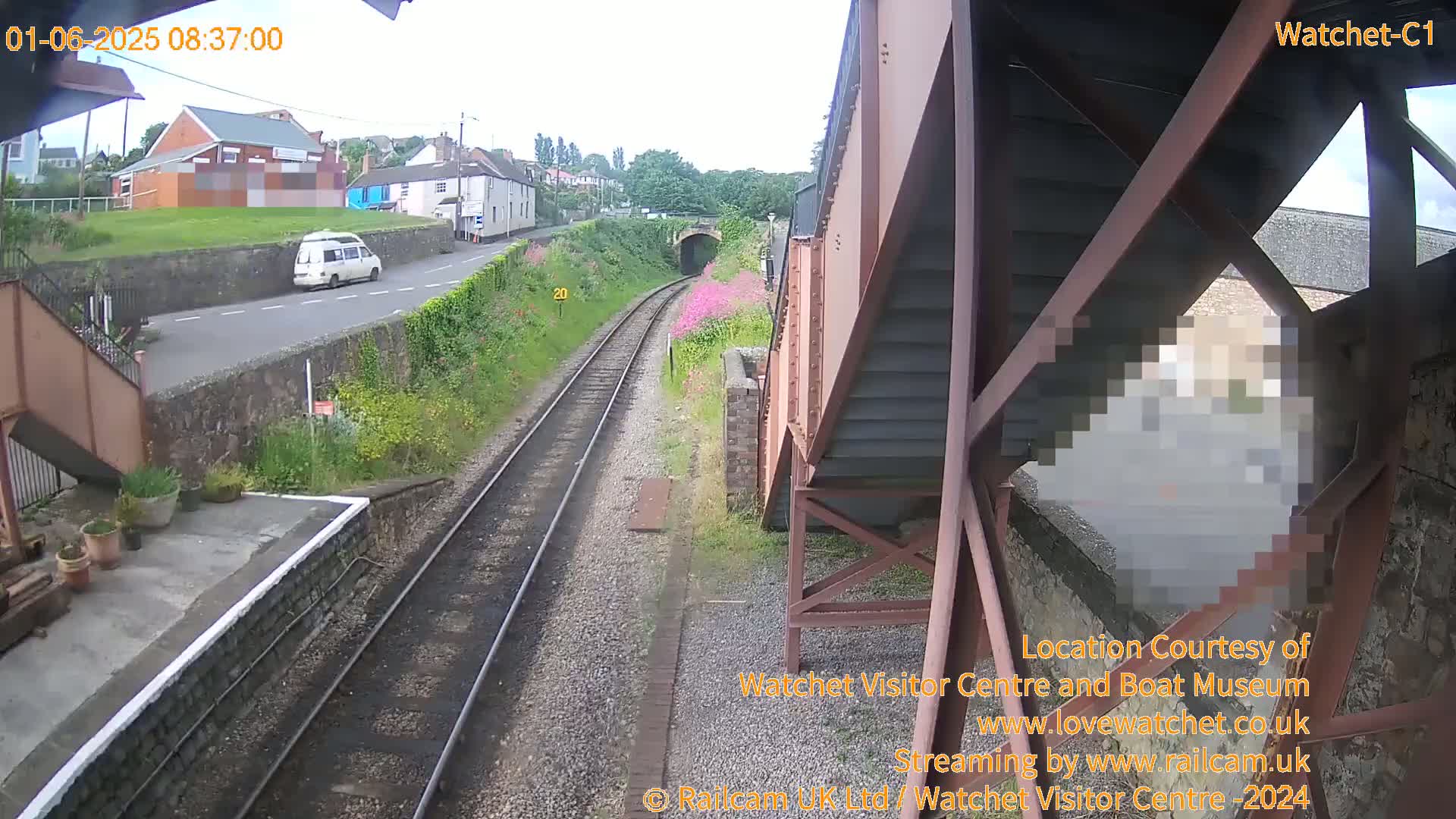 A view from a railway bridge shows a train track, a road with a white van, and buildings under an overcast sky.