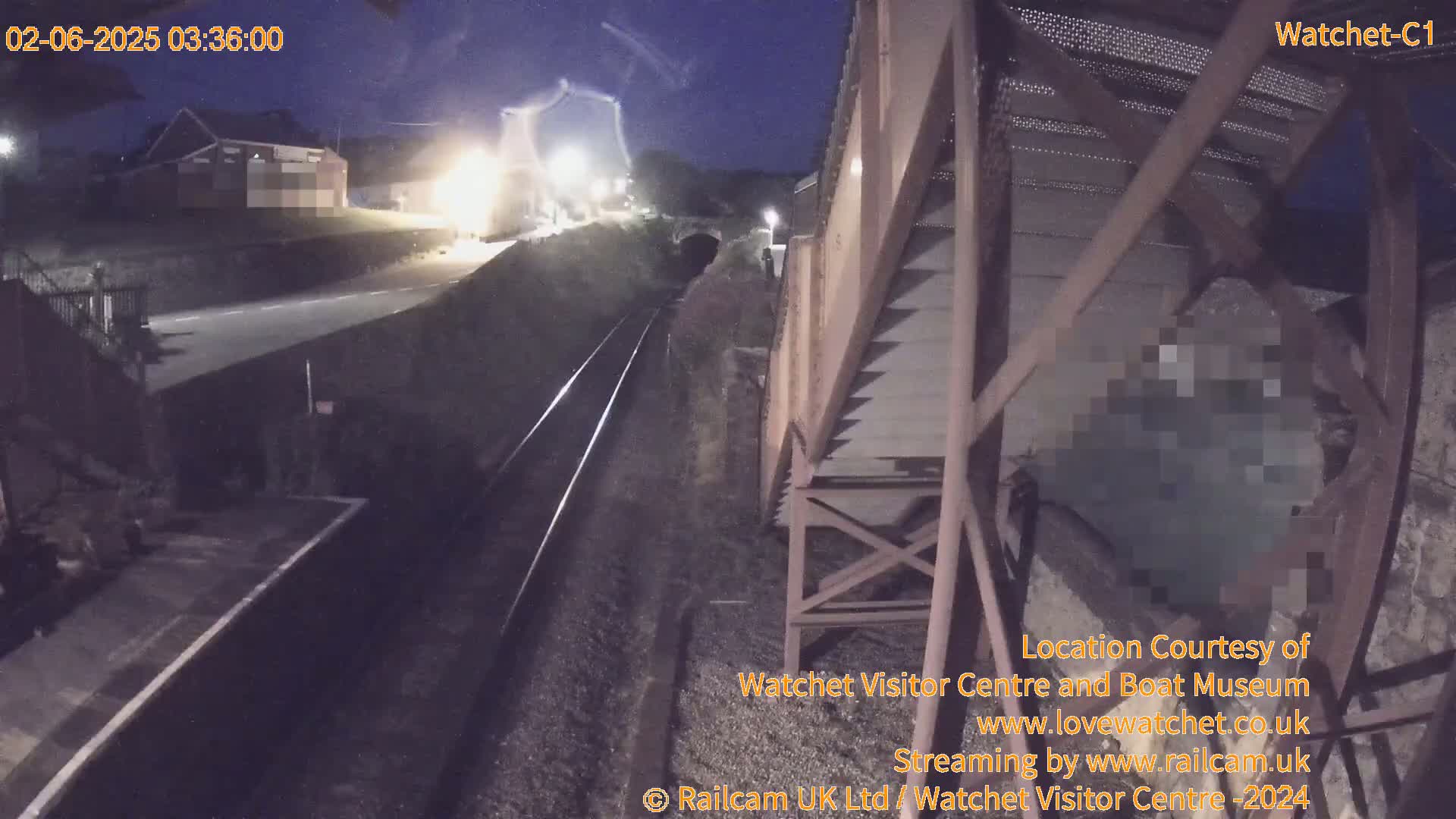 A nighttime view from a high vantage point shows a railway track extending into a tunnel, with a road and building visible in the distance under a dark sky.