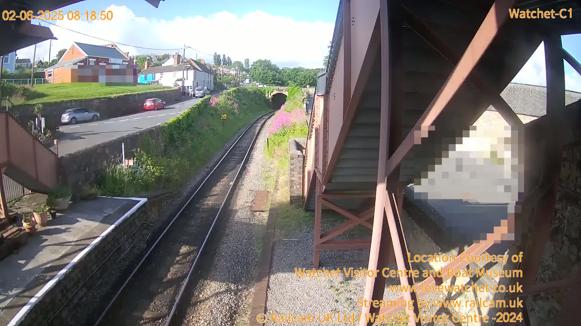 A view from a railway platform on a sunny day shows train tracks leading toward a tunnel, with cars visible on a road above, and a building in the background.
