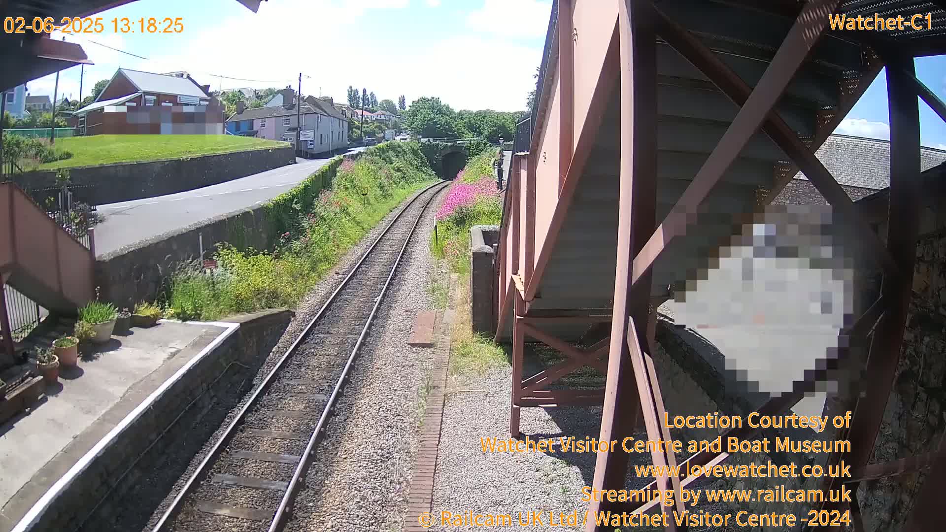 A sunny view from a railway platform shows train tracks leading to a tunnel, with a road and buildings visible beyond.