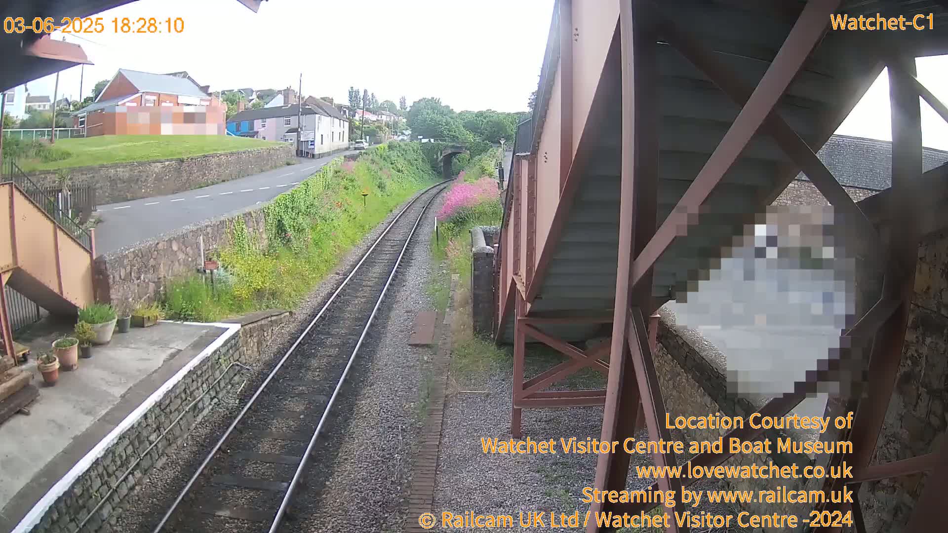 A railway track with a person visible through a metal structure, overlooked by a building and a road, under overcast skies.