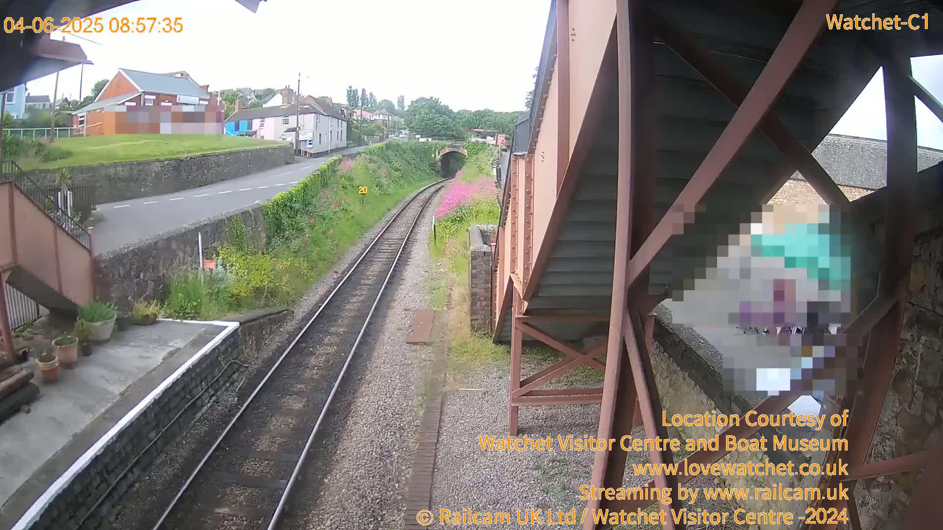 A view from a train shows a railway track leading to a tunnel, alongside a road and buildings, under an overcast sky.