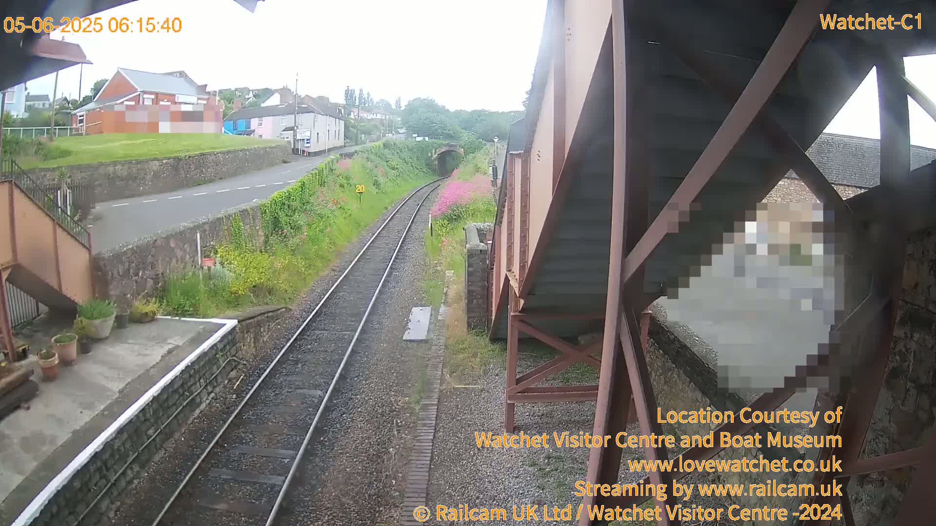 A train track extends toward a tunnel under an overcast sky, with a road and buildings visible in the background.