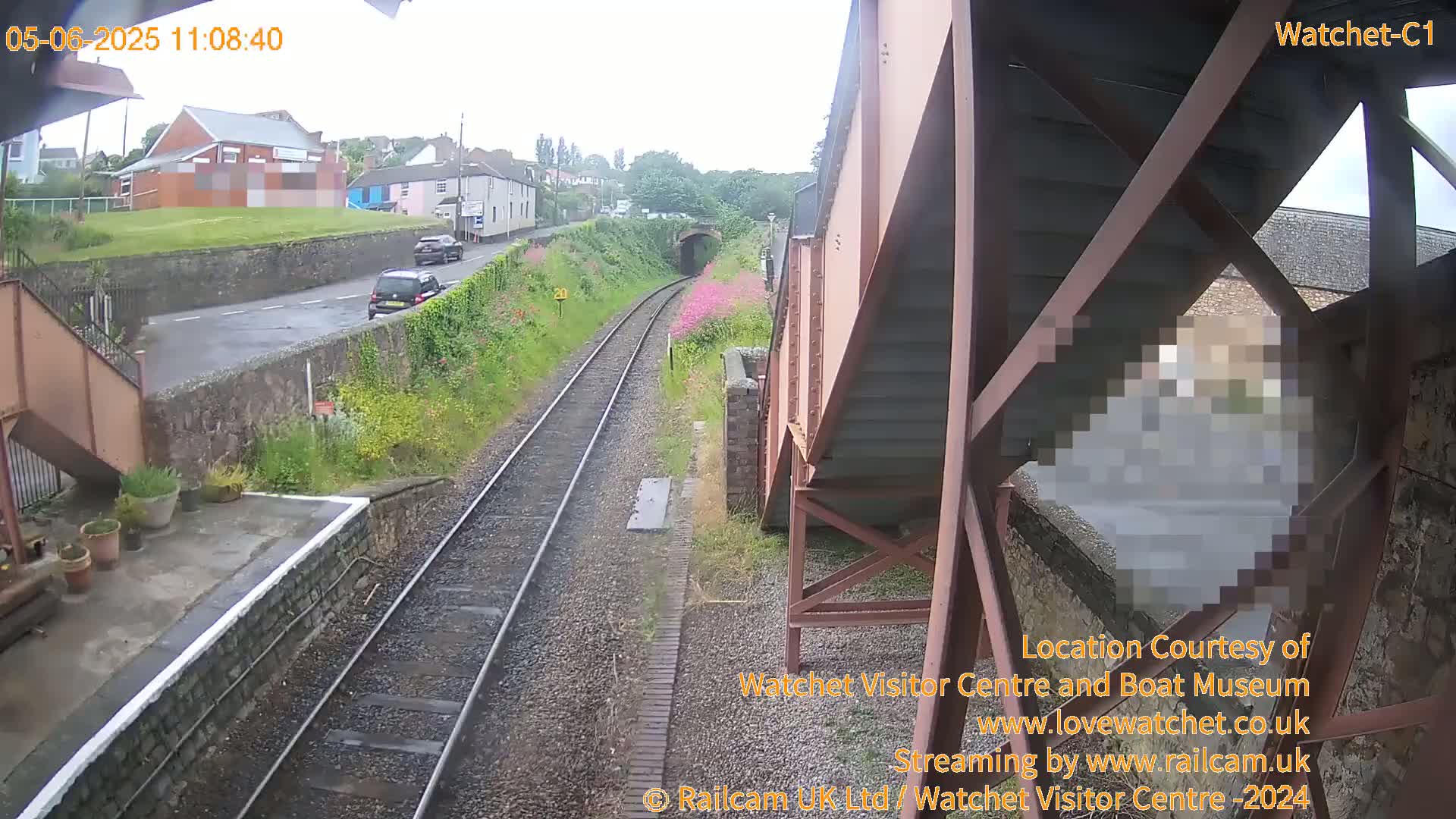 A view from a footbridge shows a railway line with two cars on a road next to it, under an overcast sky.