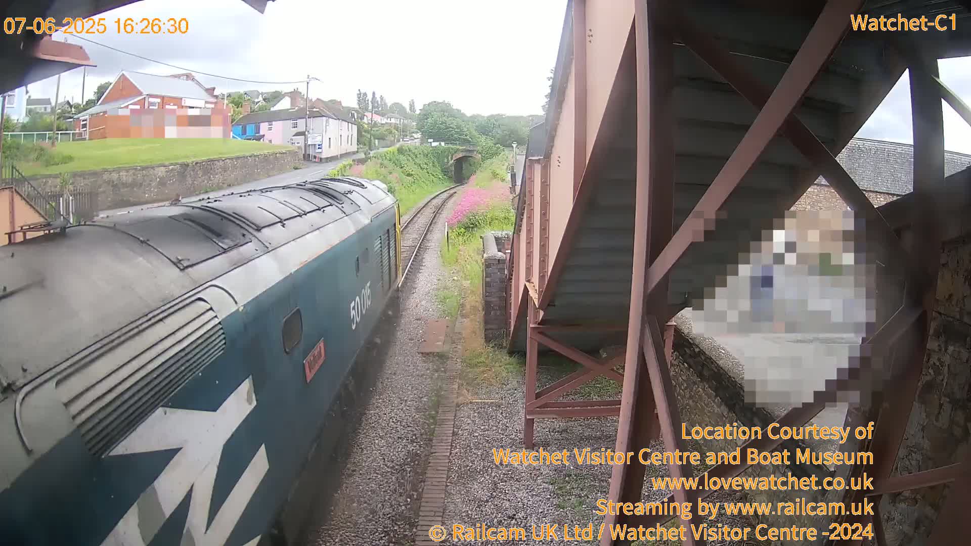 A blue and white diesel locomotive approaches a railway tunnel under a metal footbridge on a rainy day.