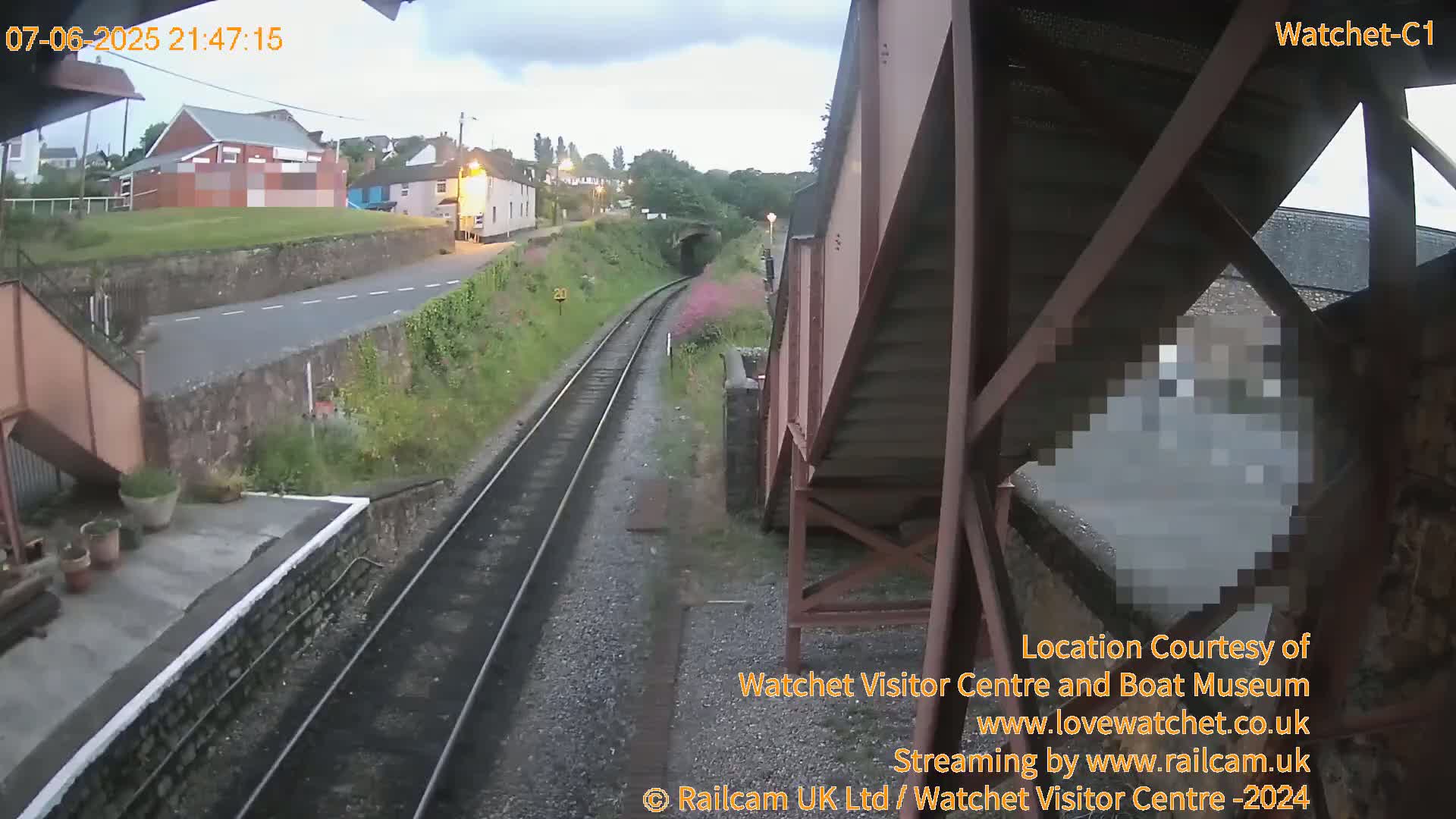 A view from a railway footbridge at dusk shows train tracks leading to a tunnel, with buildings and a road visible in the distance.