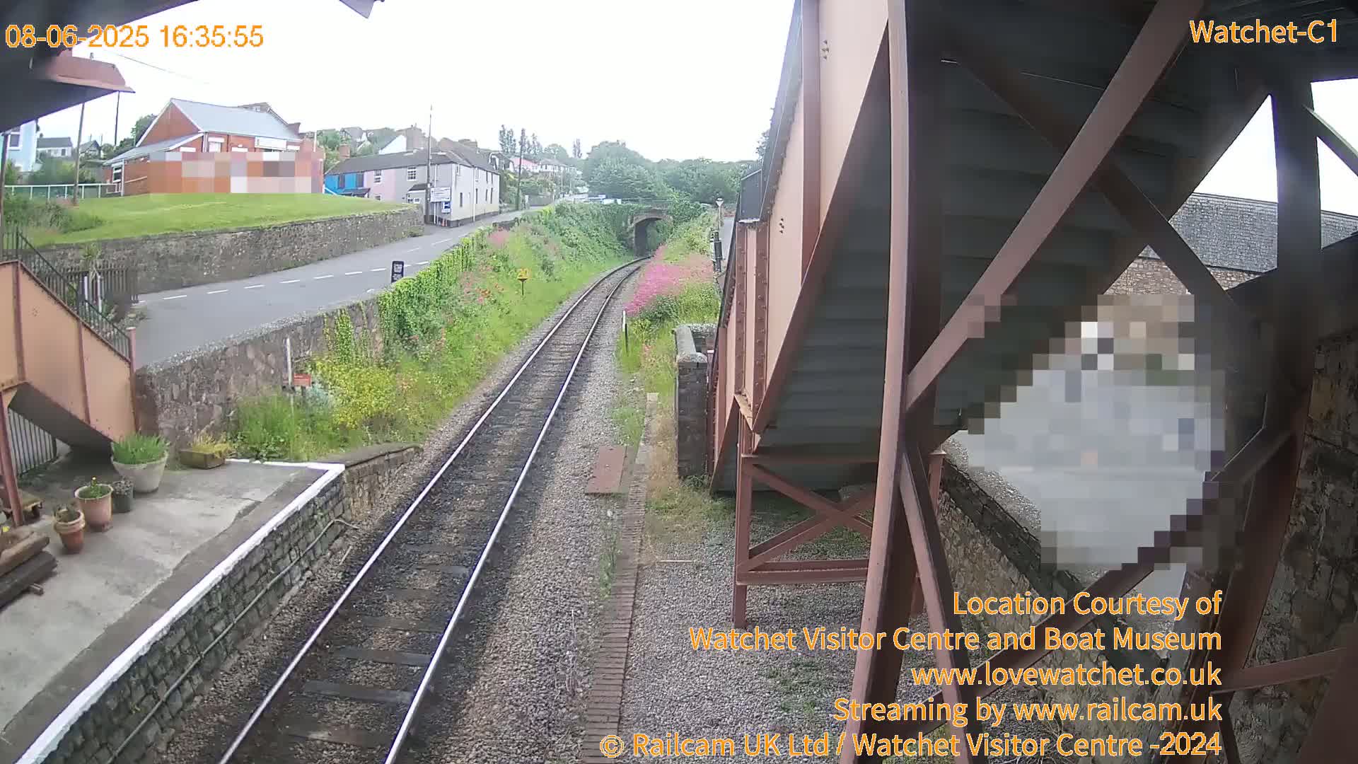 A view from under a metal footbridge shows train tracks leading towards a tunnel and a small town under an overcast sky.