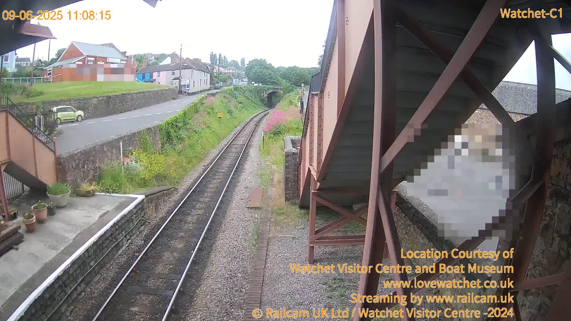 A view from a train trestle shows a railway line leading to a tunnel, a road with a small car, and buildings in the background on an overcast day.