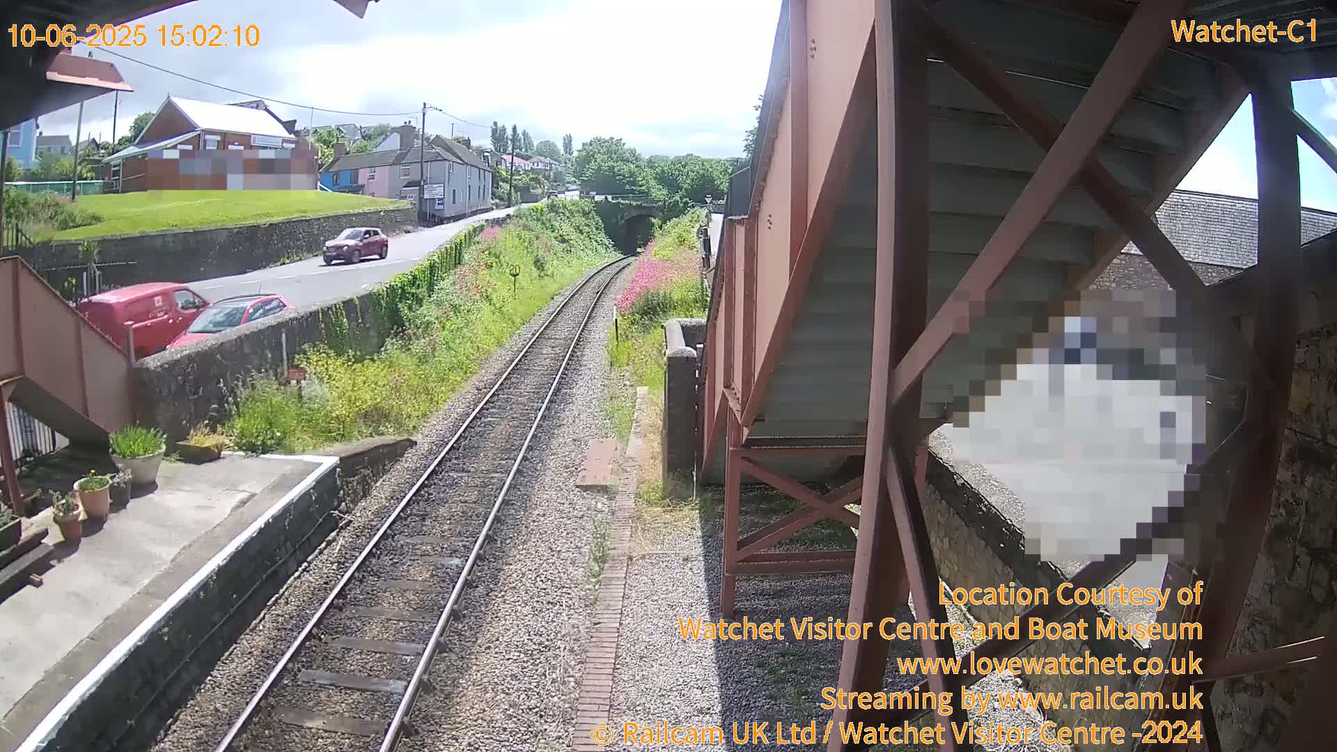 A railway line stretches towards a tunnel under a sunny sky, with a road visible alongside it and several cars driving by.
