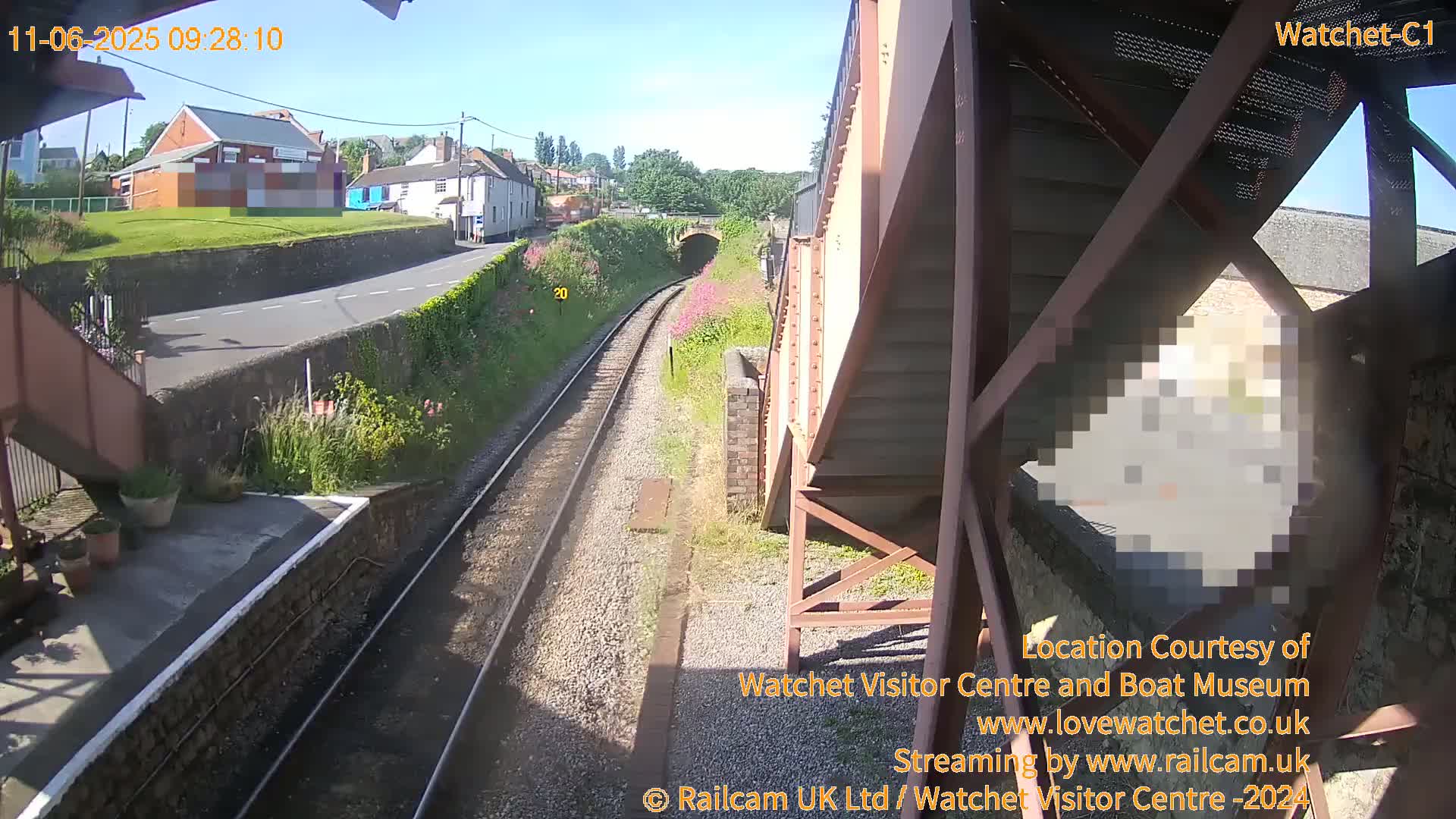 A sunny view from a railway platform shows train tracks leading to a tunnel, with a road and buildings visible in the distance.