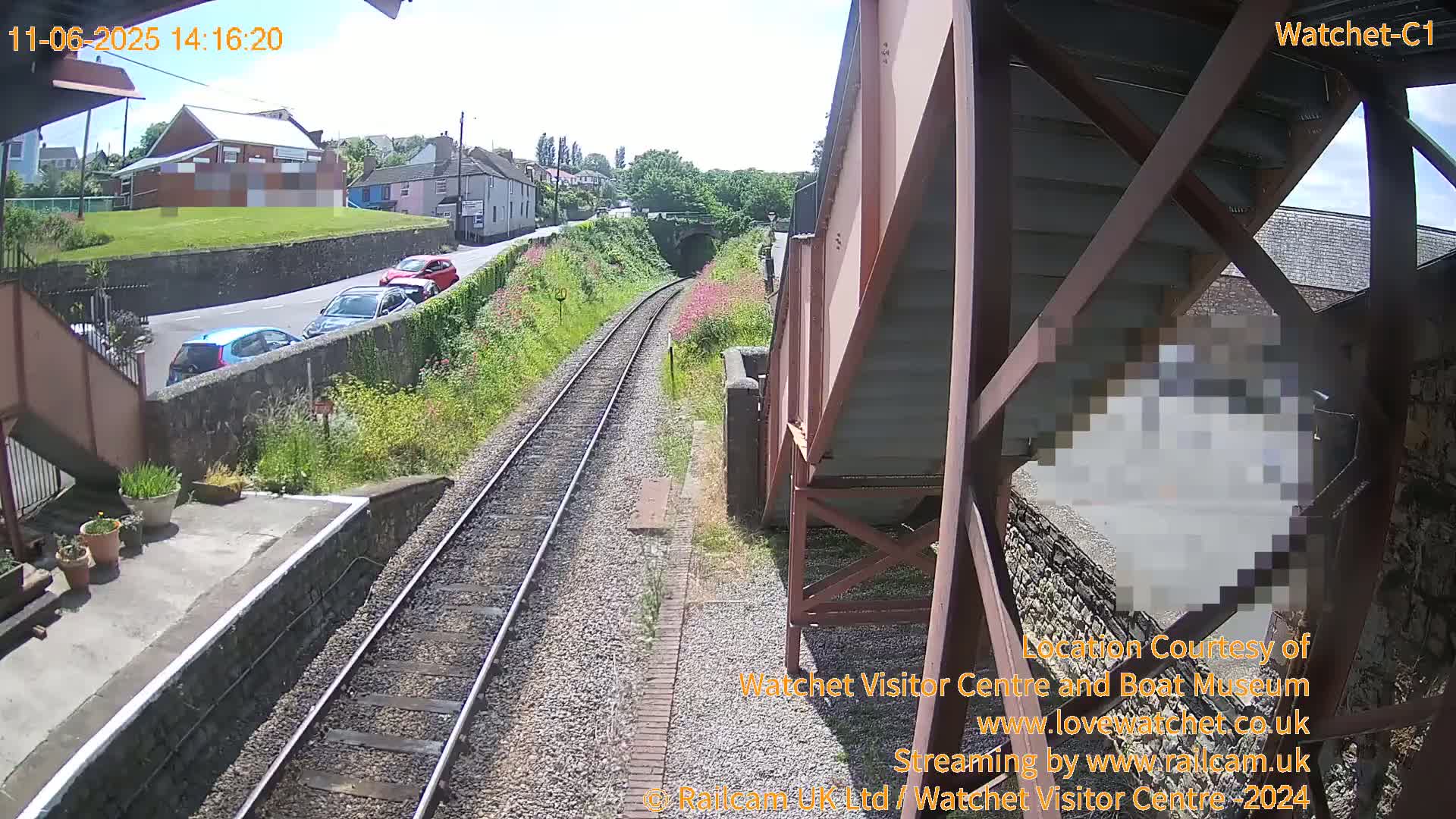 A sunny view from a railway platform shows train tracks leading to a tunnel, with several cars parked along a road beside a grassy hill and buildings in the background.