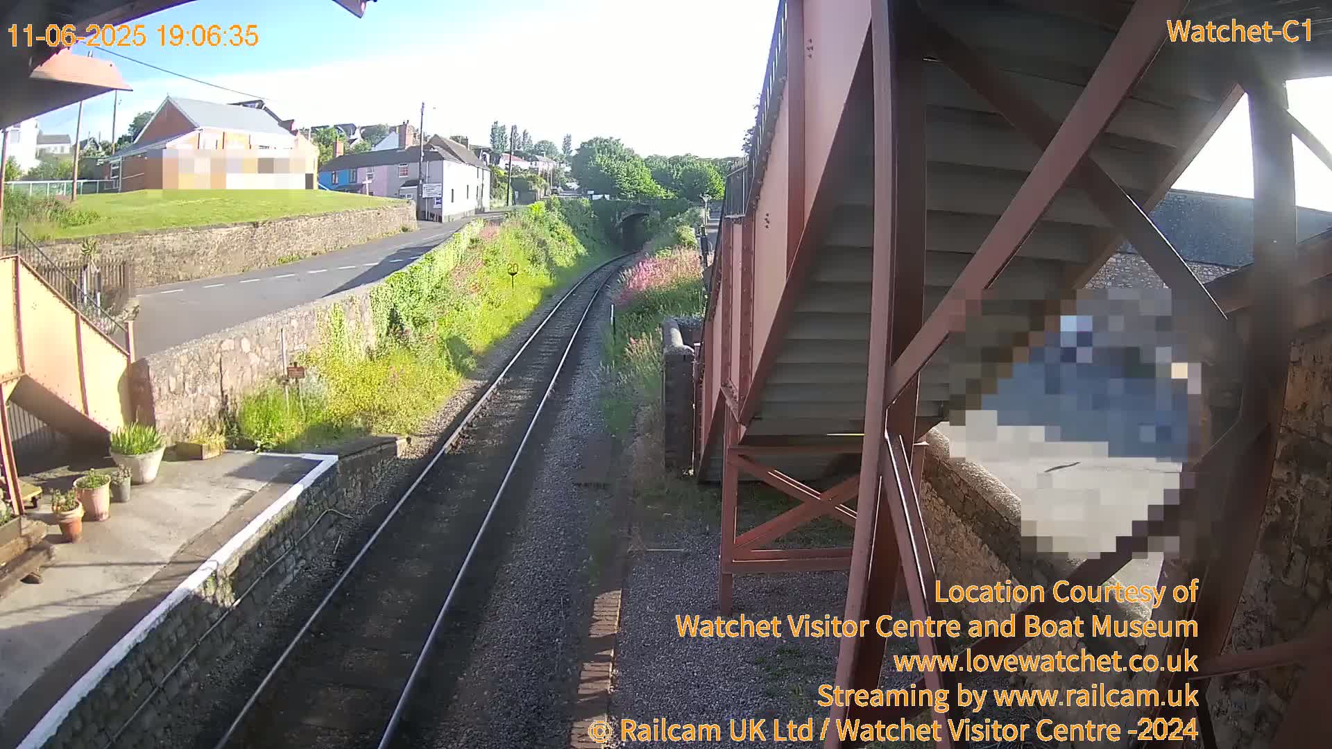 A view from a railway footbridge on a sunny evening shows train tracks leading towards a small town with houses and a stone wall, alongside a road.