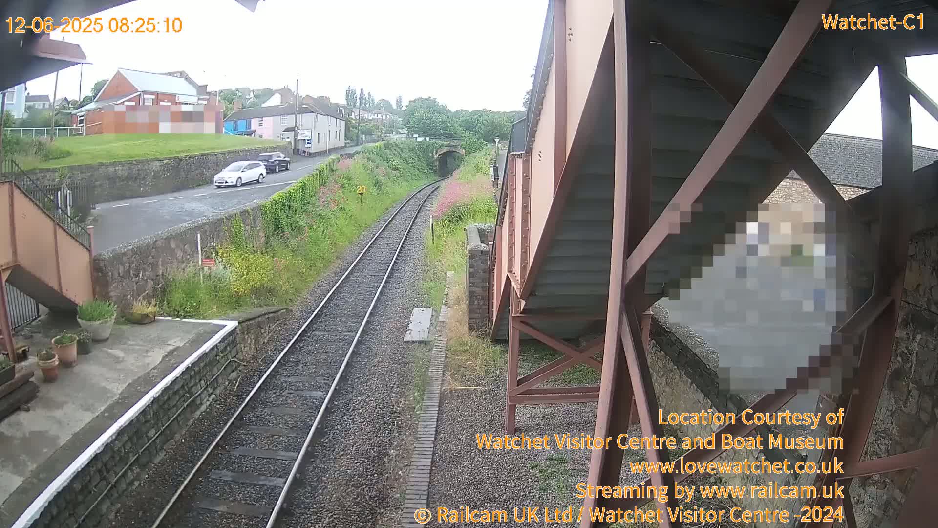 A view from a train shows a railway track leading to a tunnel, a road with two cars, and a building, all under an overcast sky.
