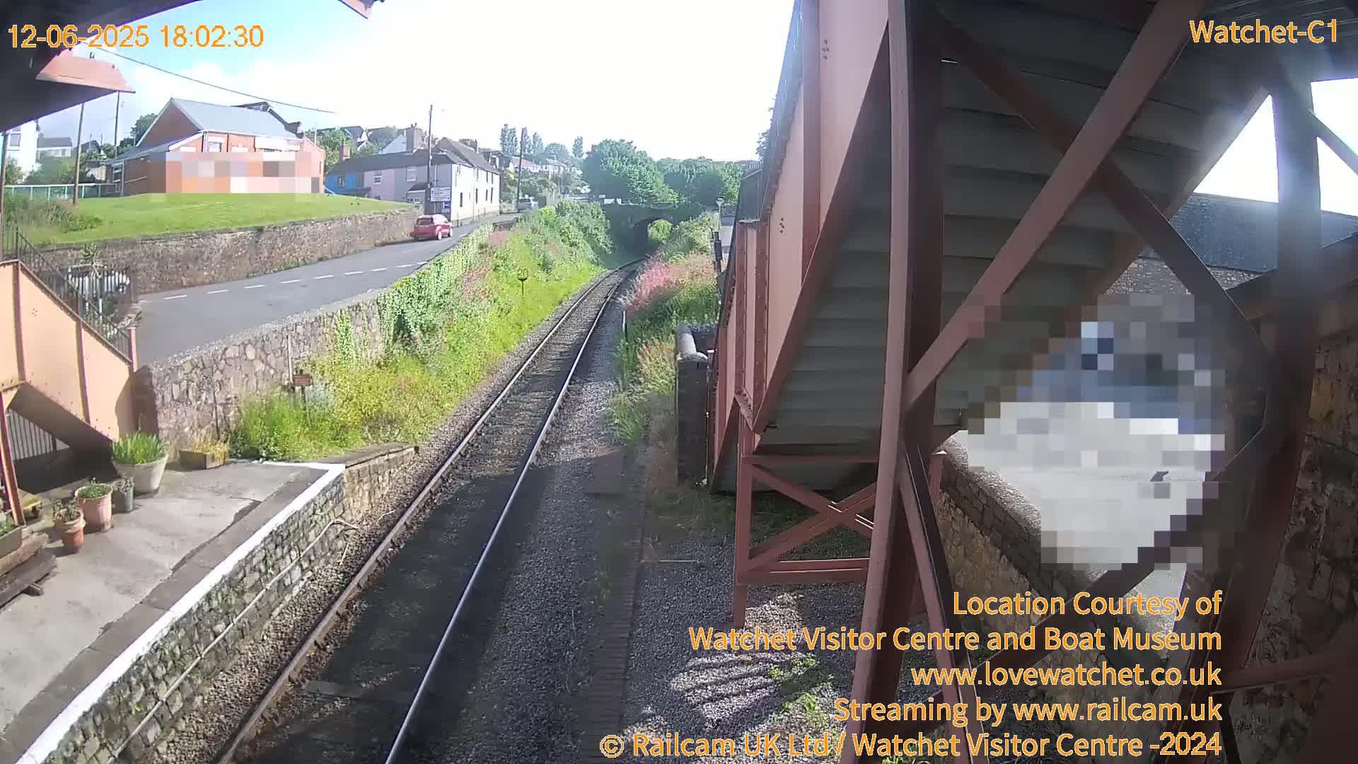 A sunny view from a train station platform shows train tracks extending into the distance, a road with a car, and buildings in the background.