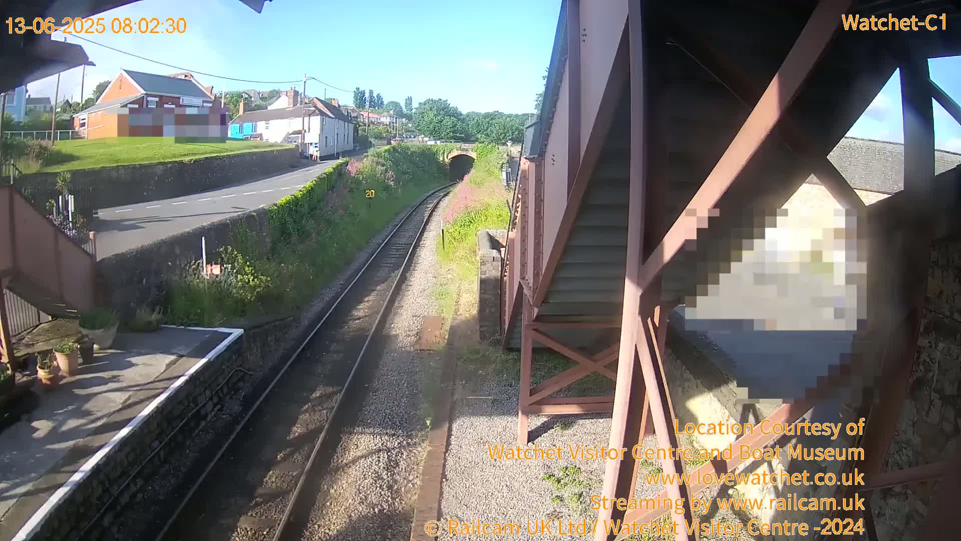 A sunny view from a railway platform shows train tracks leading to a tunnel, with a road and buildings visible beyond.