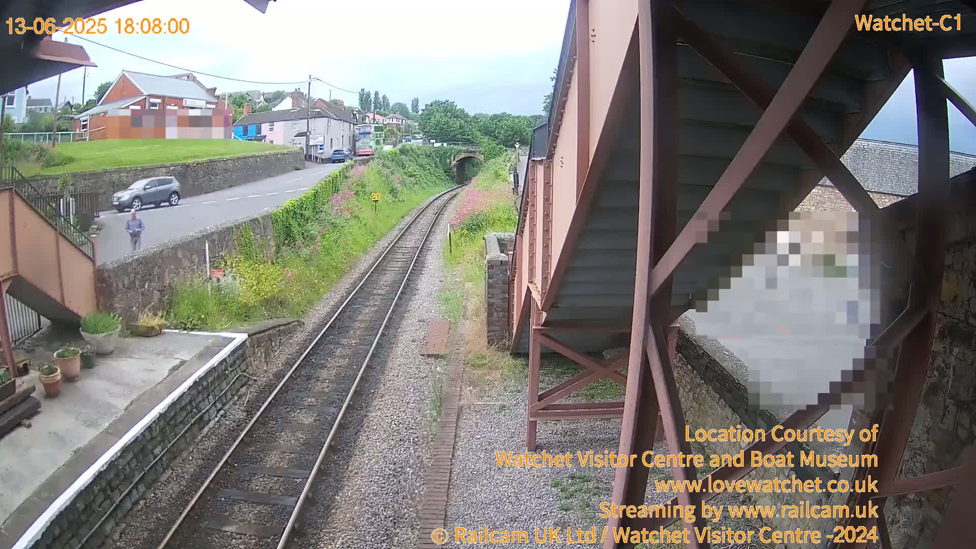 A view from a train platform shows a railway line, a road with a car and pedestrian, and buildings in the background on a partly cloudy day.