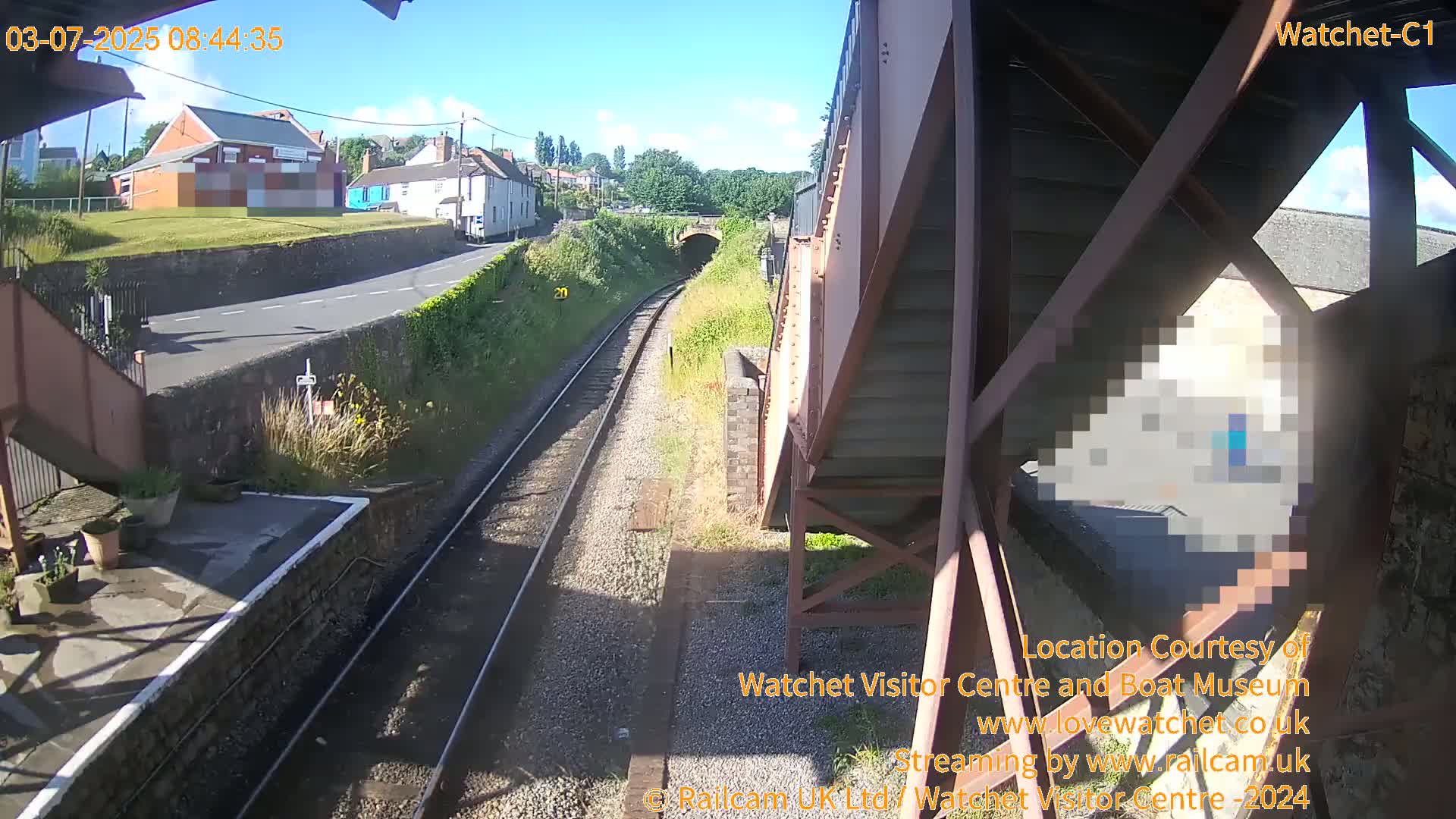 A sunny view from a train platform shows railway tracks leading to a tunnel, with a road and buildings visible in the distance.