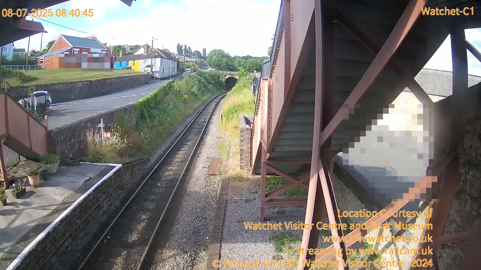 A sunny view from a railway footbridge shows train tracks leading to a tunnel, a road with a car, and buildings in the distance.