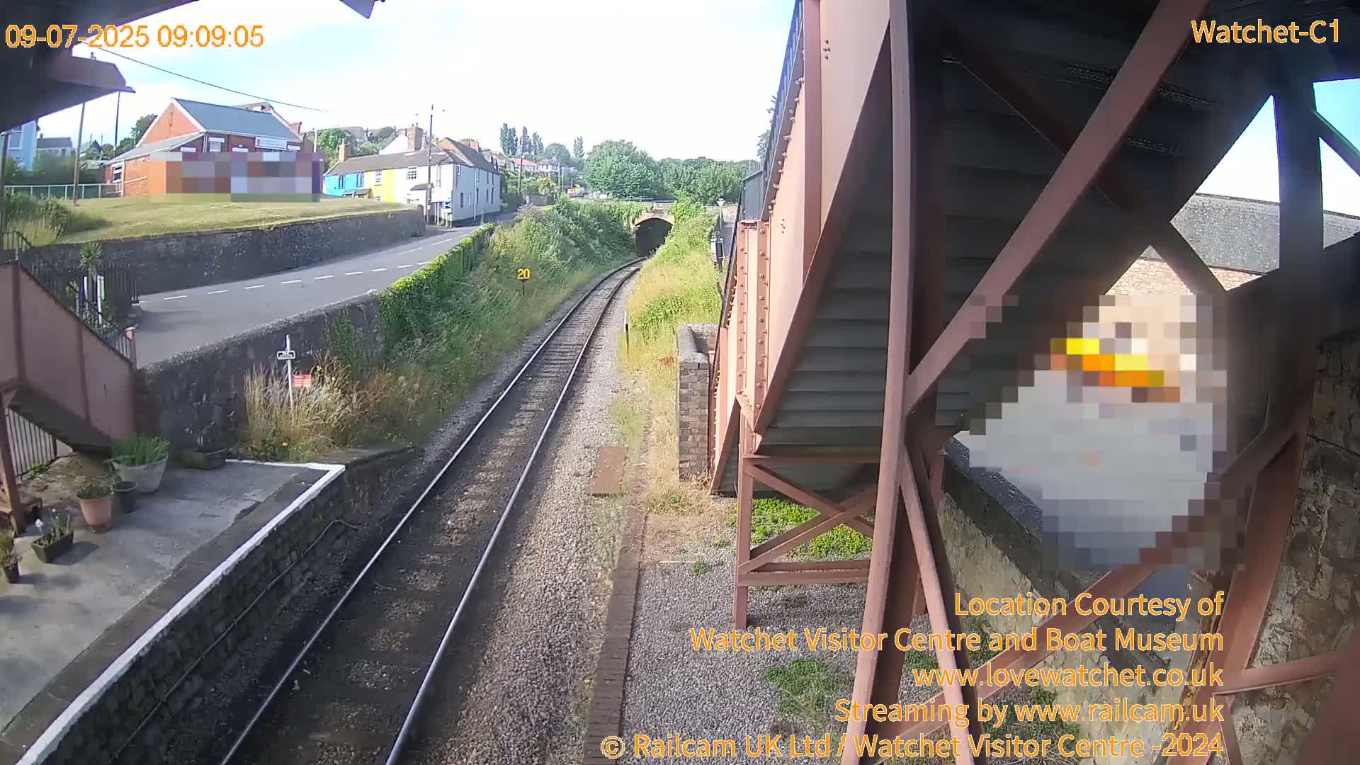 A view from under a railway bridge on a sunny day shows railway tracks leading to a tunnel, with a road and buildings visible in the distance.