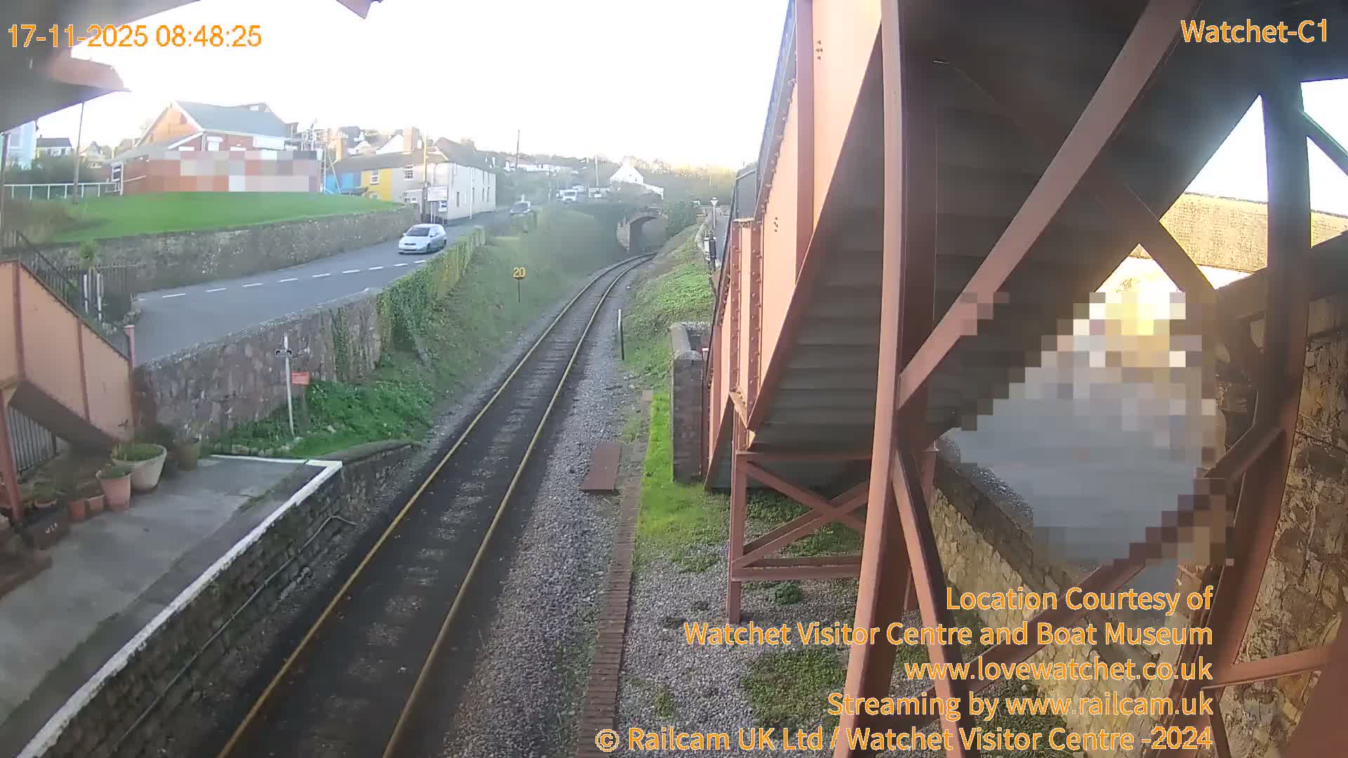 On a bright, clear morning, a railway line curves past a station platform and alongside a road with a white car and distant buildings, with a large reddish-brown metal structure prominently on the right.