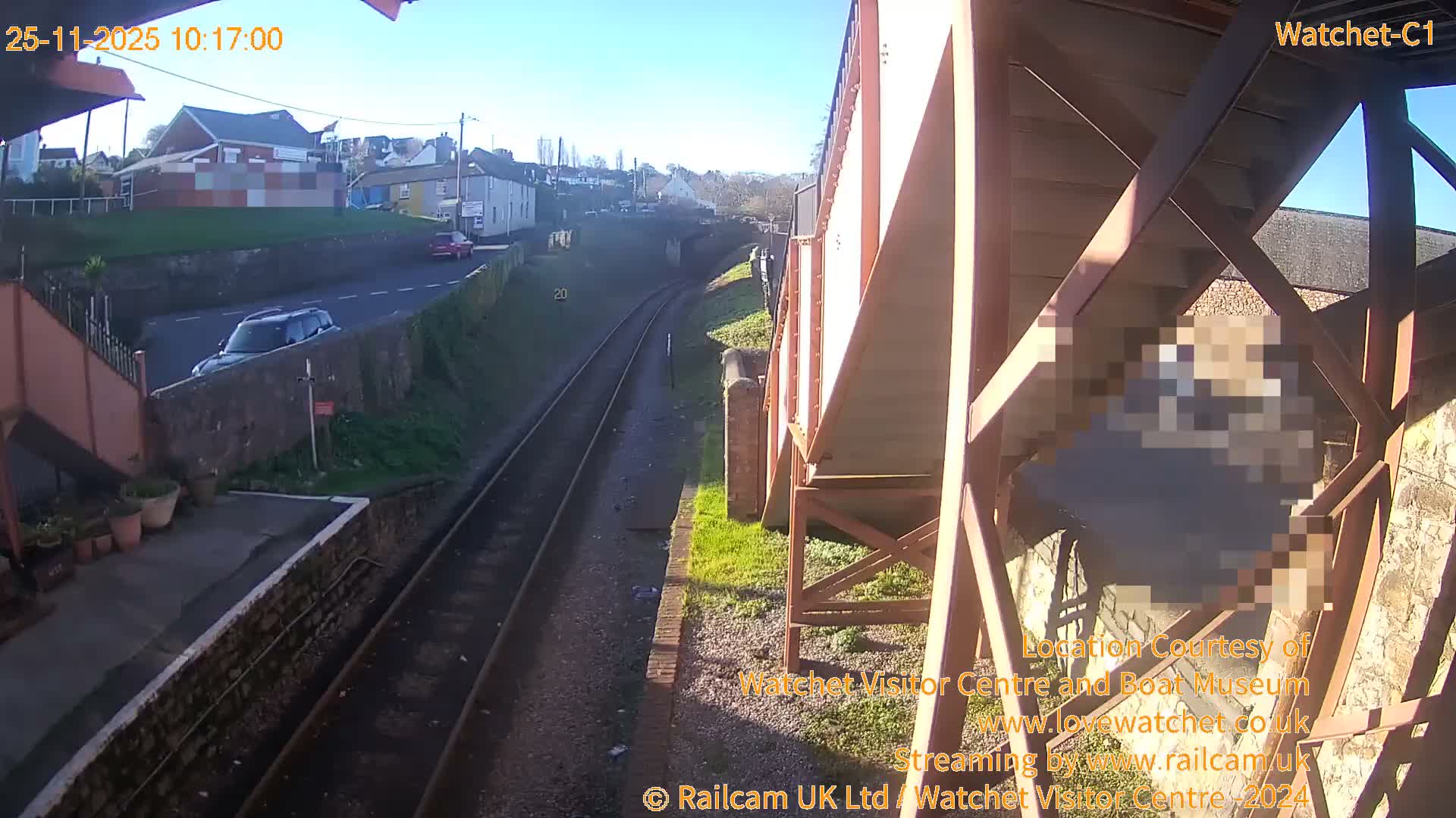 This outdoor image displays train tracks curving through a sunny landscape with a platform on the left, a road with cars above, and buildings on a distant hill, all viewed from under a large metal bridge structure on a clear day.
