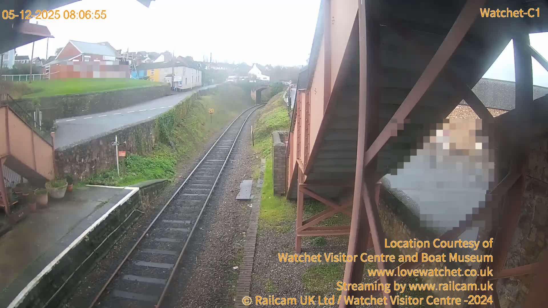 On an overcast and wet day, a train track extends into the distance through a town, bordered by a station platform on the left and partially by a reddish pedestrian bridge on the right, with a road and houses visible on the higher ground.