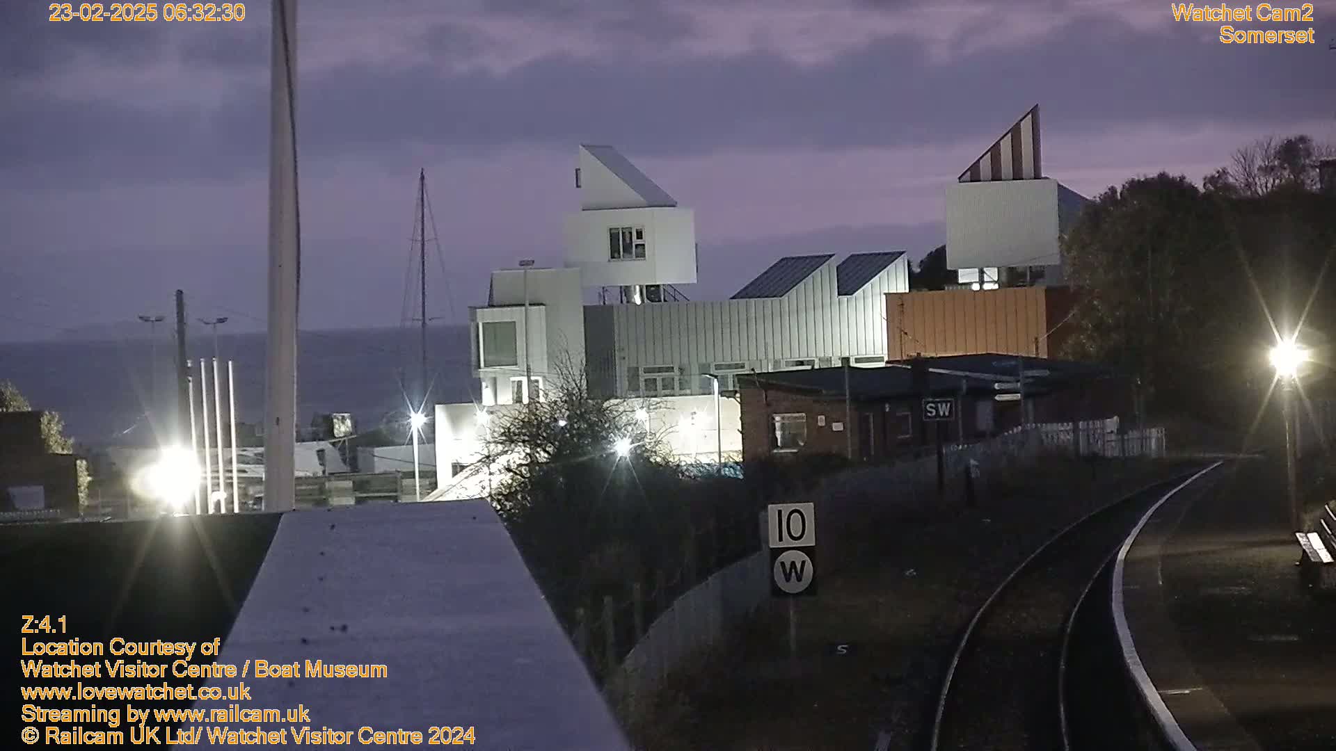A modern building complex is visible near a body of water under a dusky, pre-dawn sky, with train tracks in the foreground.