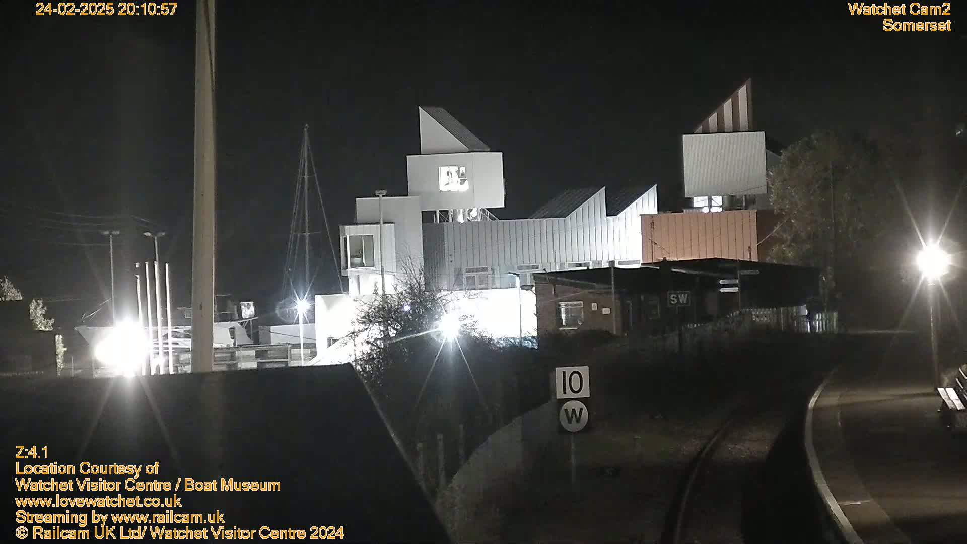 At night, a modern, angular building complex is illuminated, next to a railway line with a speed limit sign.