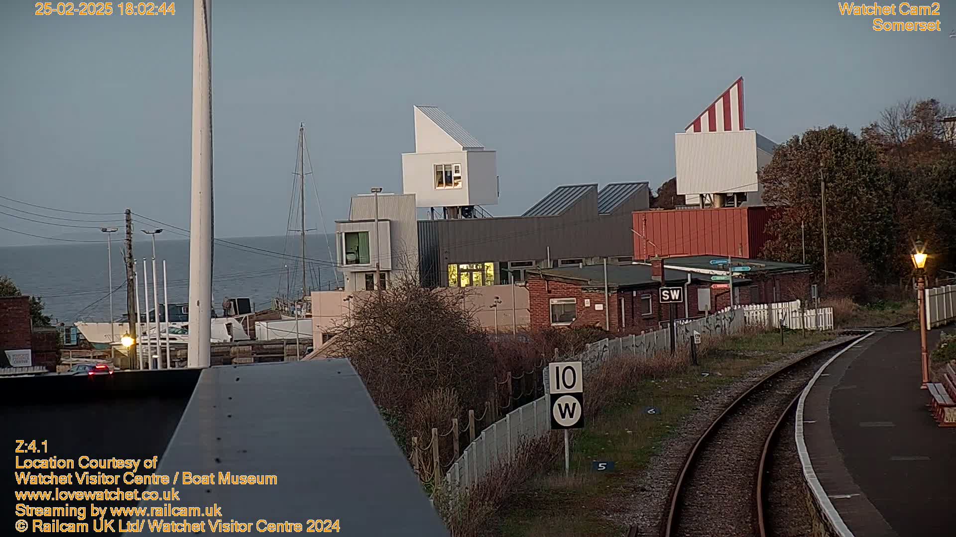 A seaside town scene at dusk shows modern buildings near a train track and body of water.