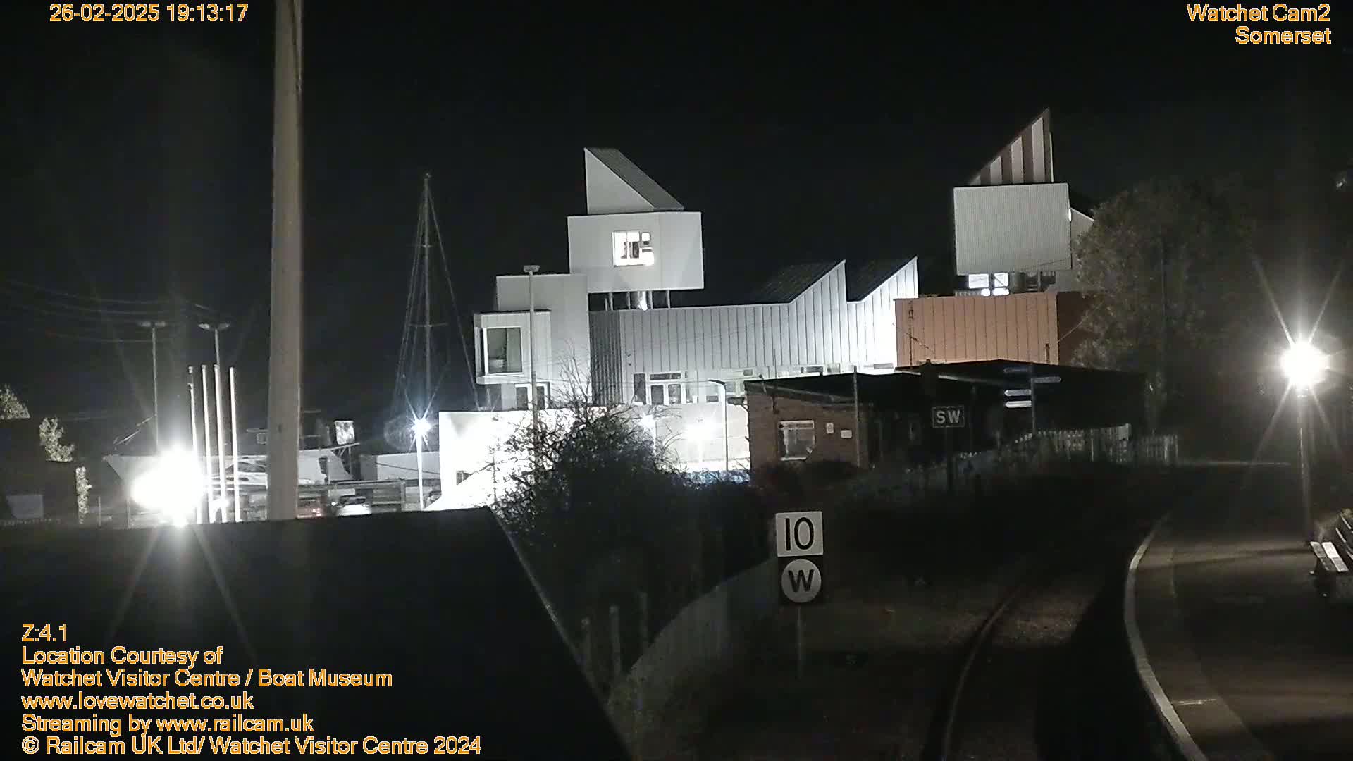 At night, a modern white building complex is illuminated, situated near a railway line and some trees.