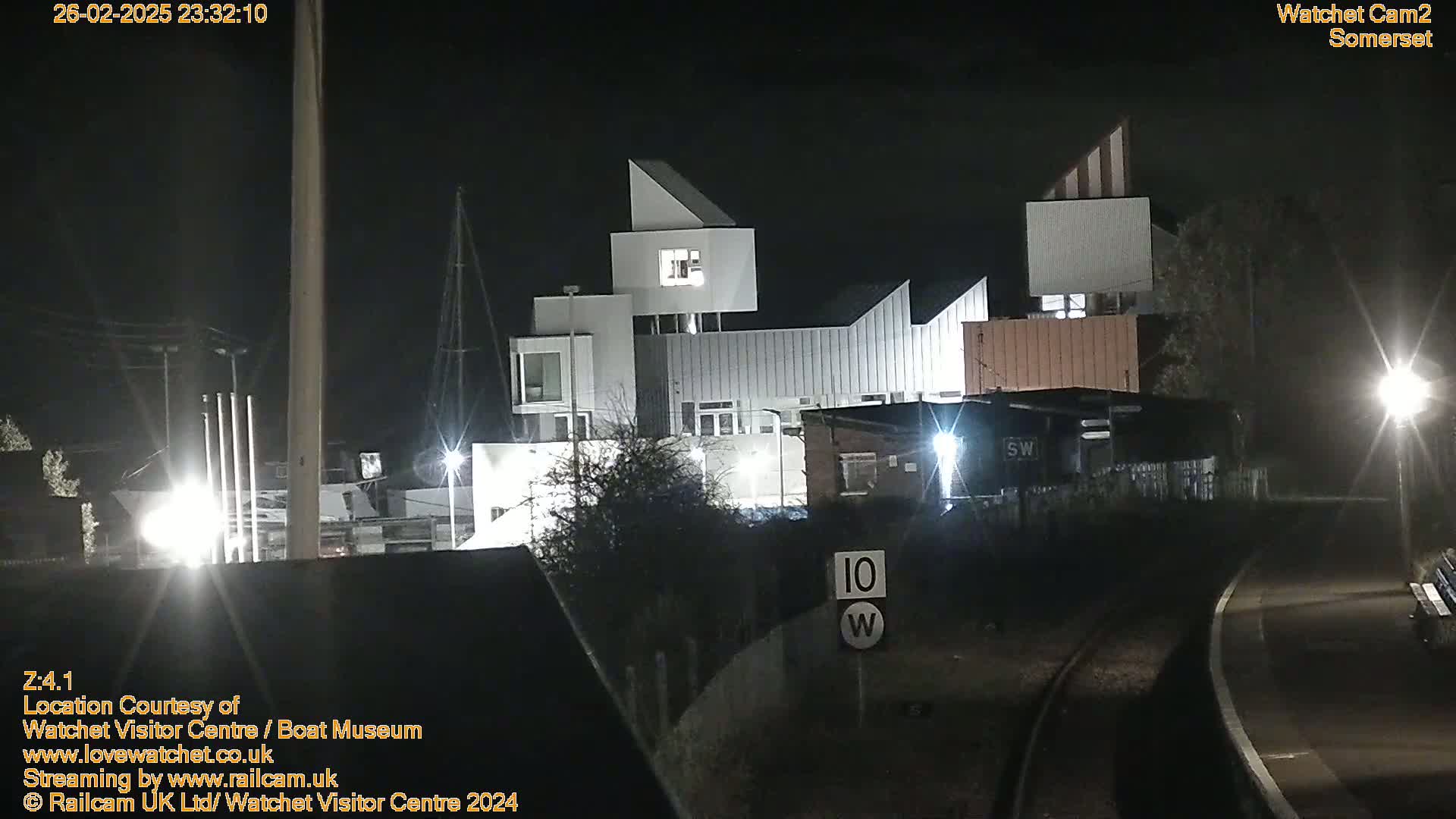 At night, a modern building complex is illuminated, near railway tracks and a speed limit sign.