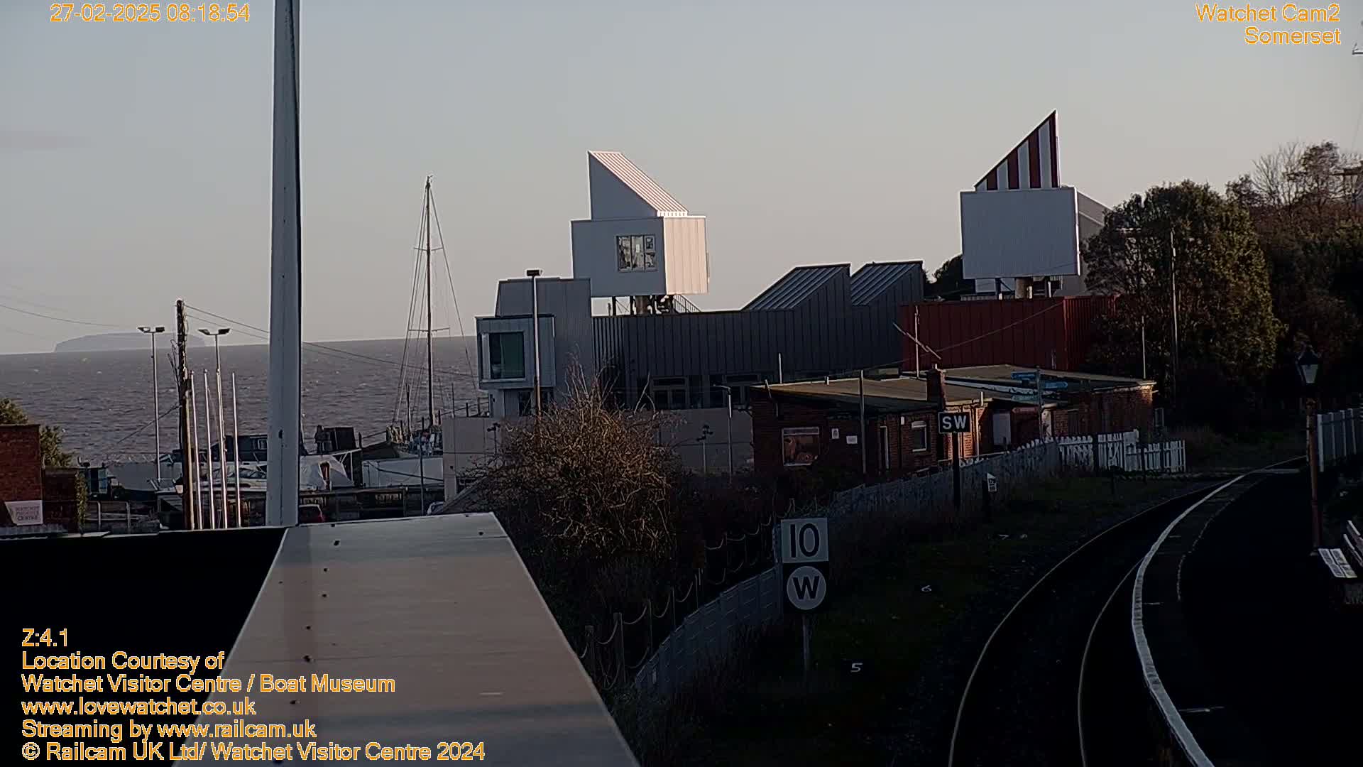 A waterfront view shows modern buildings near train tracks under a clear, dusky sky.