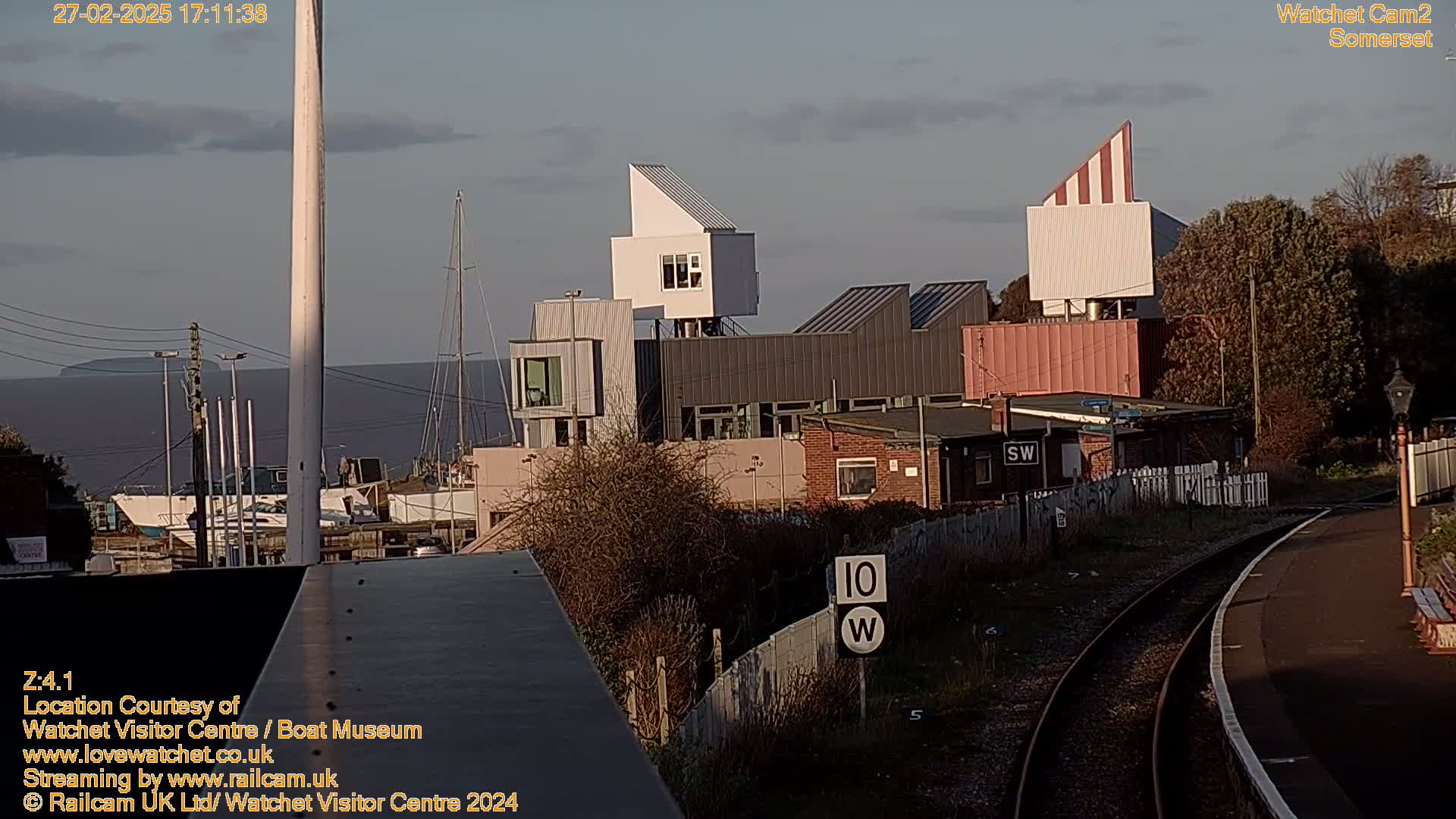 A waterfront scene at sunset shows modern buildings near a railway line, with boats in a harbor and a calm sea in the background.