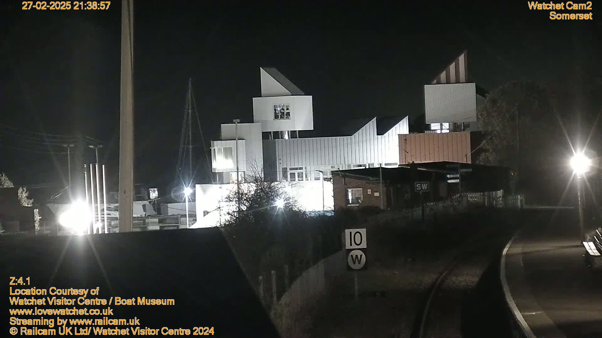A modern, multi-level building is illuminated at night, next to a railway line and road.
