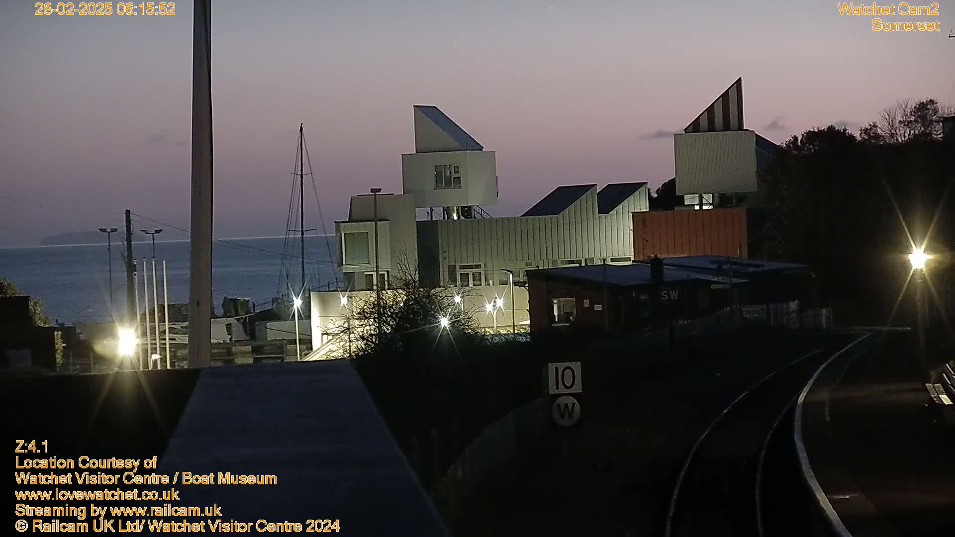A modern, multi-level building complex is illuminated at twilight near a calm body of water, with a sailboat visible in the distance.