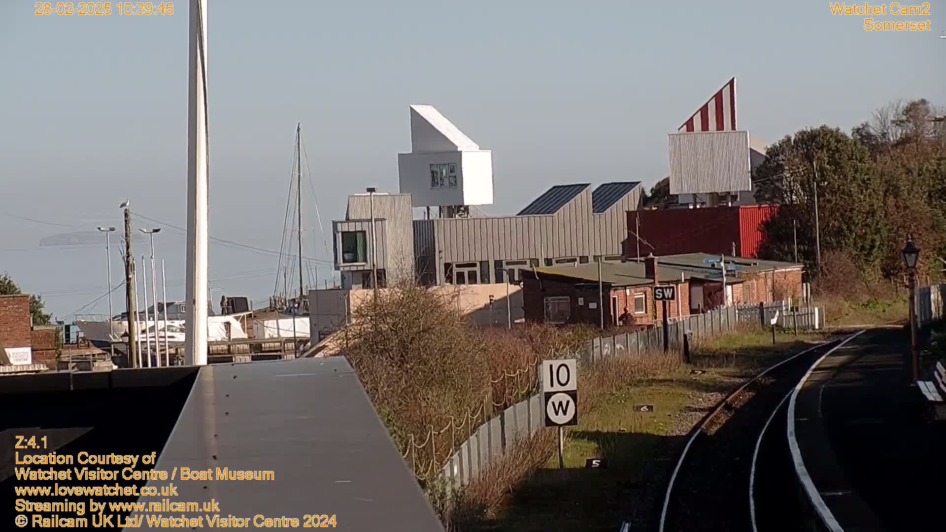 A sunny day shows a waterfront view of modern buildings, a train track, and a sailboat in the distance.