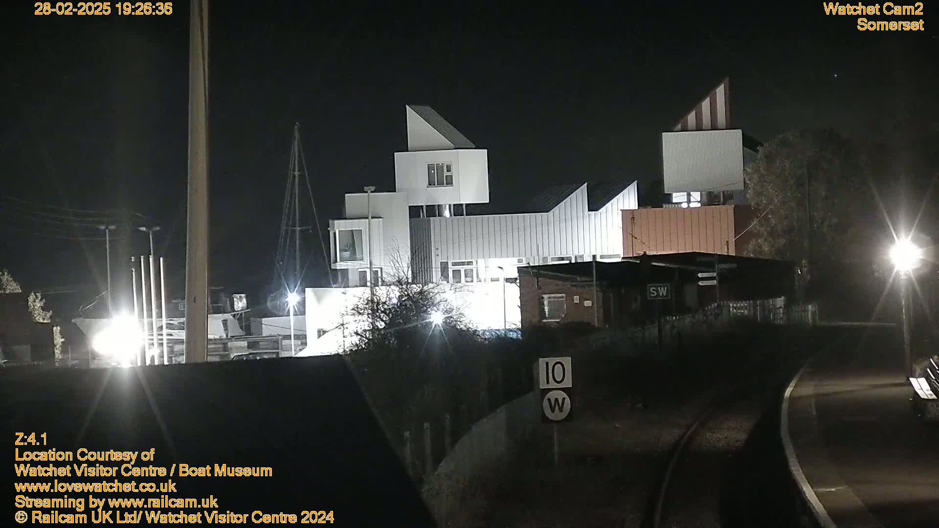 At night, a modern, multi-sectioned building is illuminated, situated near train tracks and some low shrubbery.
