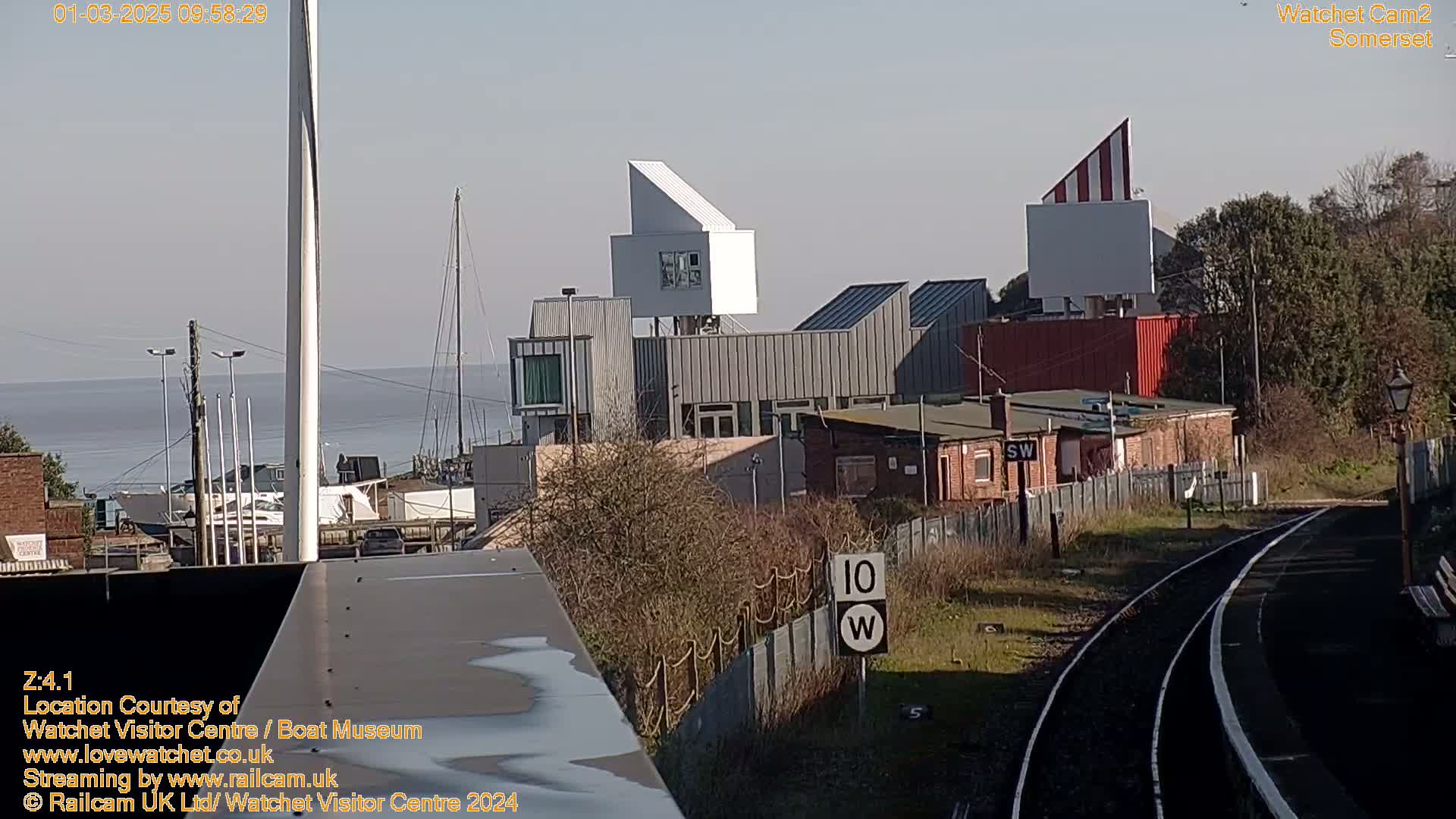 A waterfront view of modern buildings near train tracks on a sunny day, with a body of water and sailboats in the background.