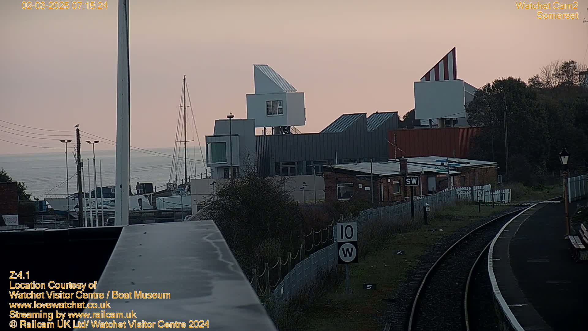 A view of a harbor with boats and modern buildings at dawn under a pale pink sky, overlooking railroad tracks.