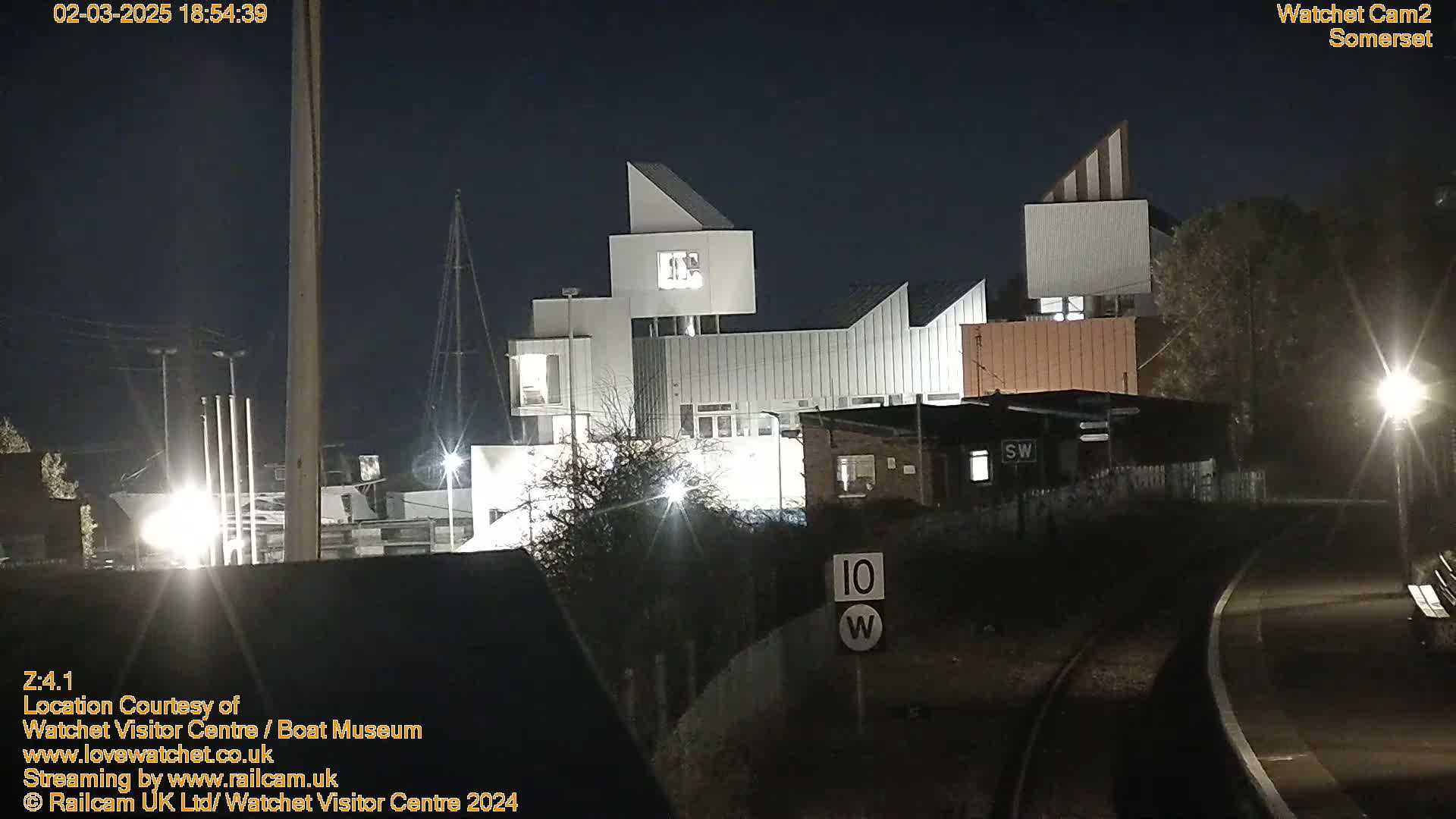 At night, a modern, multi-level building is illuminated, situated near a railway track with a speed limit sign visible.