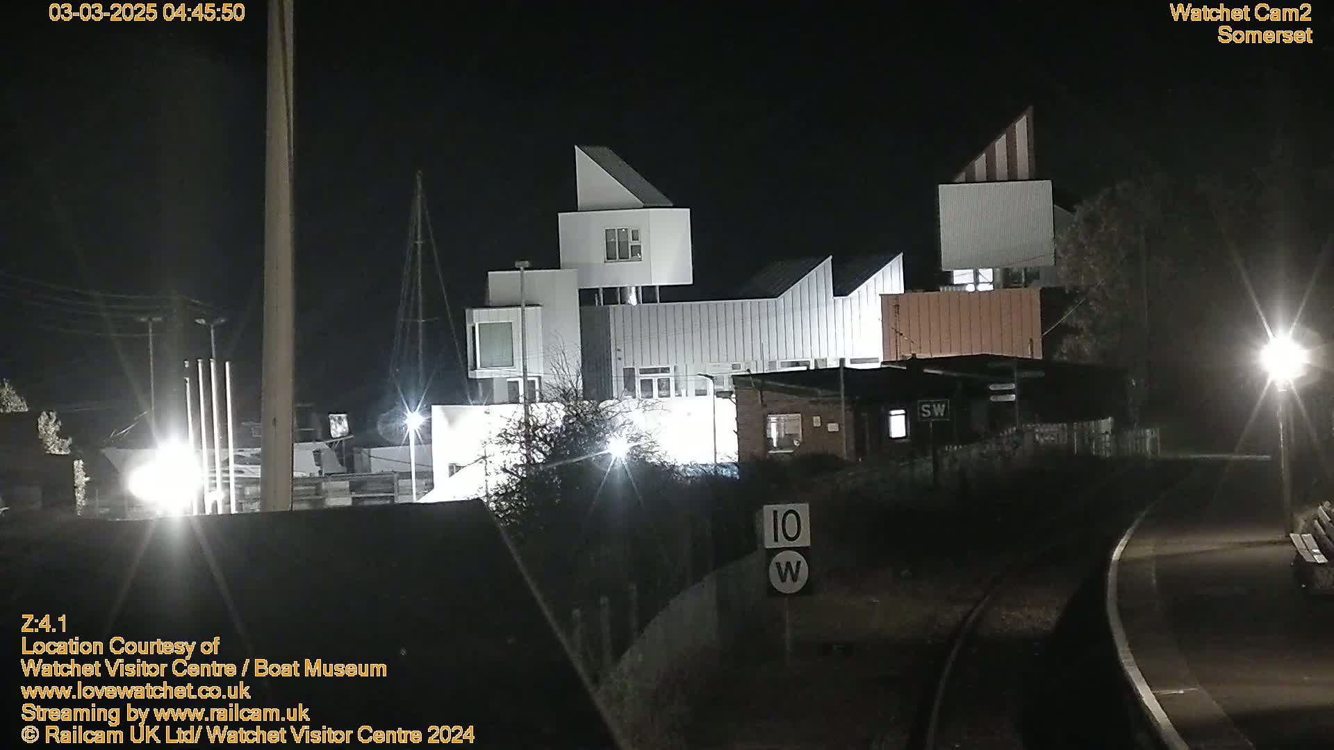 At night, a modern, multi-sectioned building is illuminated, situated near a railway track.