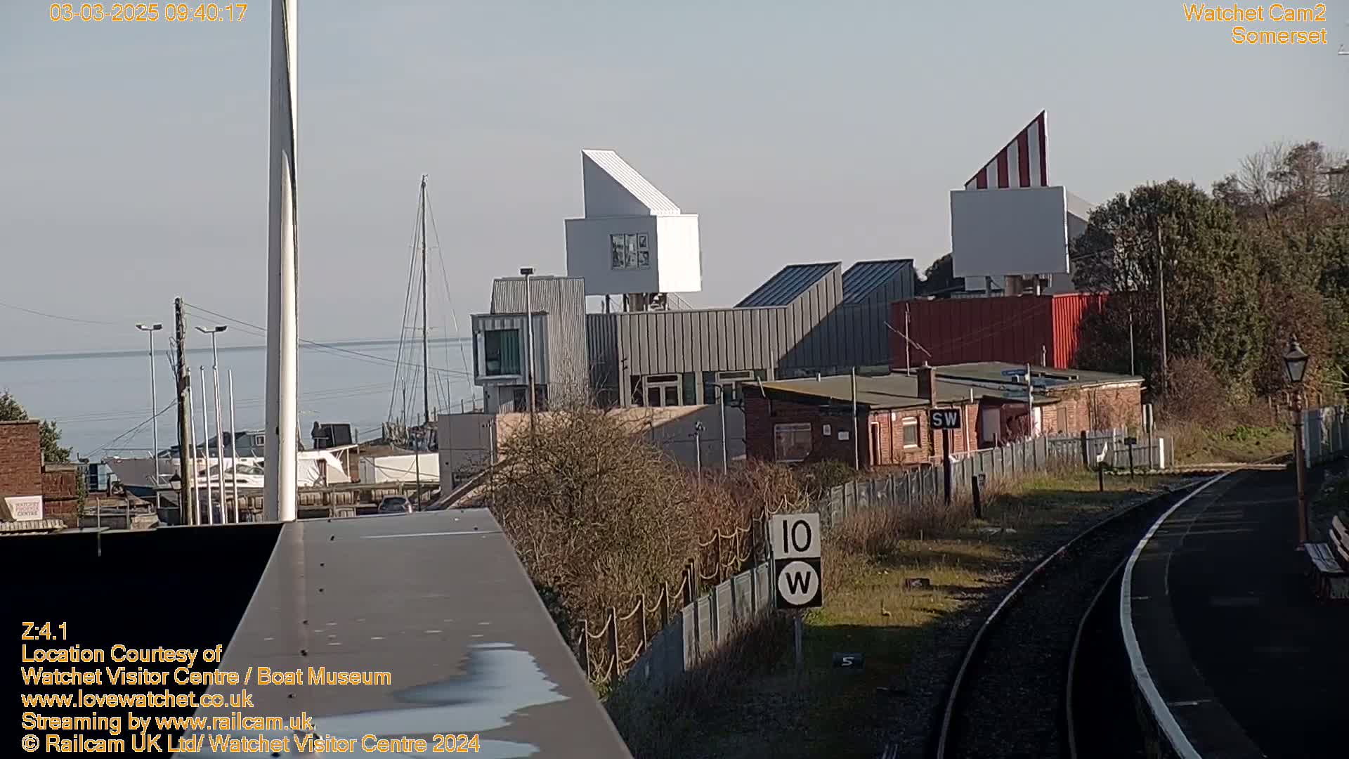 A sunny day shows a waterfront view of buildings, including a modern structure with a distinctive angular design, a railway line, and a calm body of water with boats in the distance.