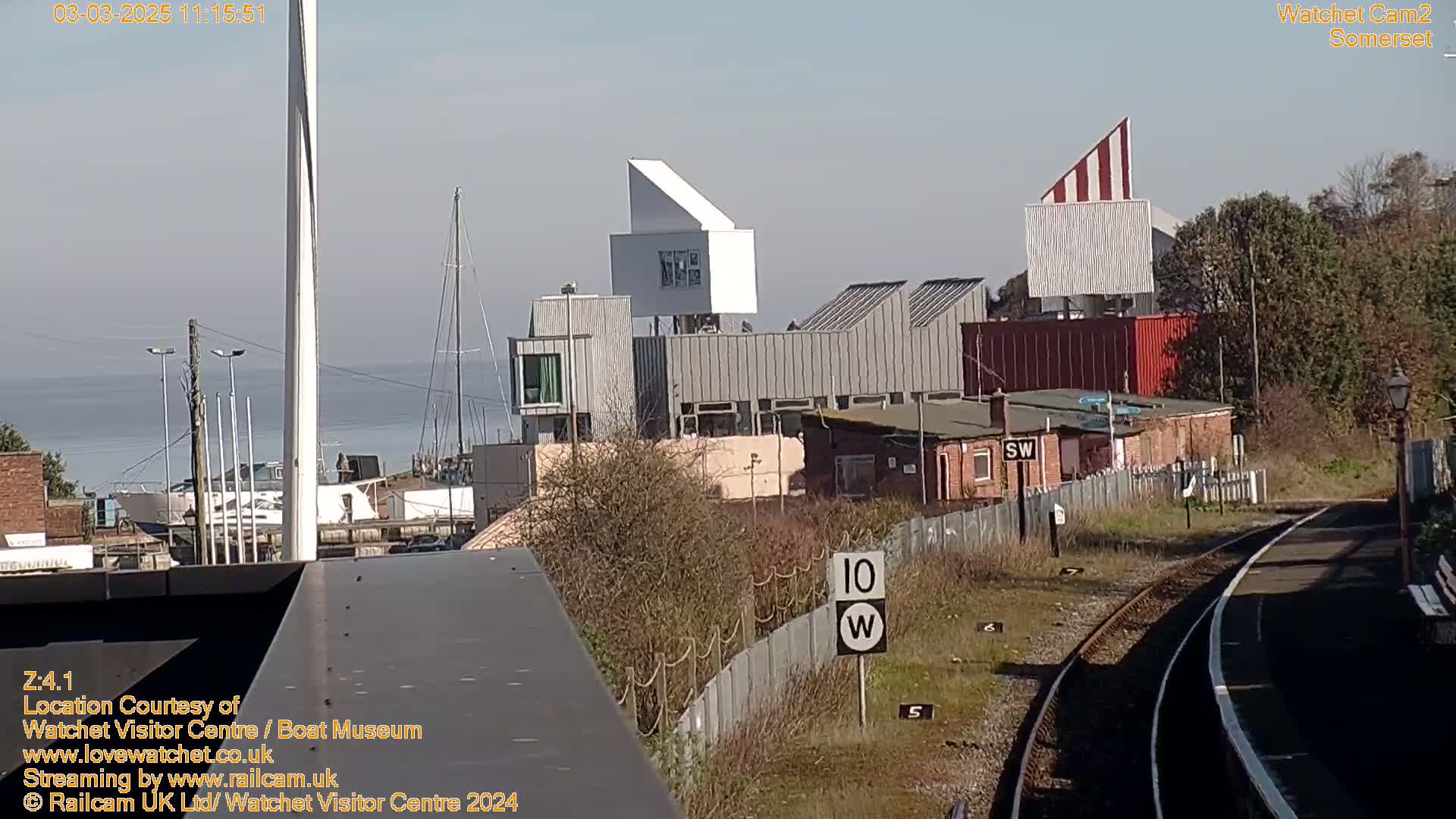 A view of a waterfront area with modern buildings, a body of water, and a railway track under a clear sky.