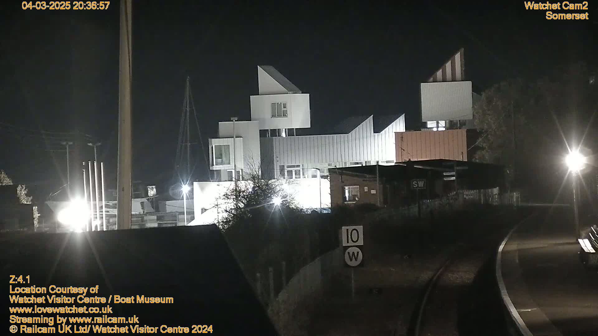At night, a modern white building complex is illuminated, next to railway tracks and a speed limit sign.