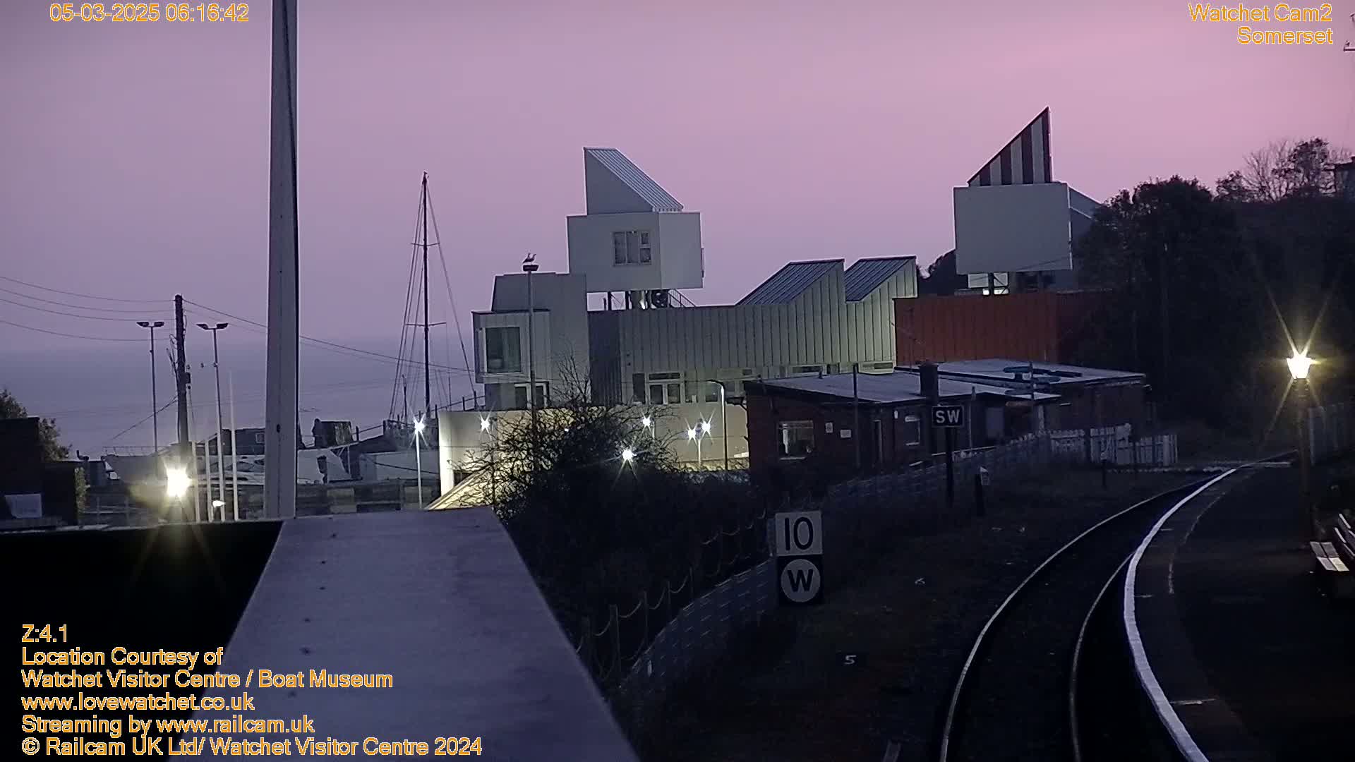 A twilight view of a waterfront area with modern buildings, a sailboat, and train tracks, under a dusky purple sky.