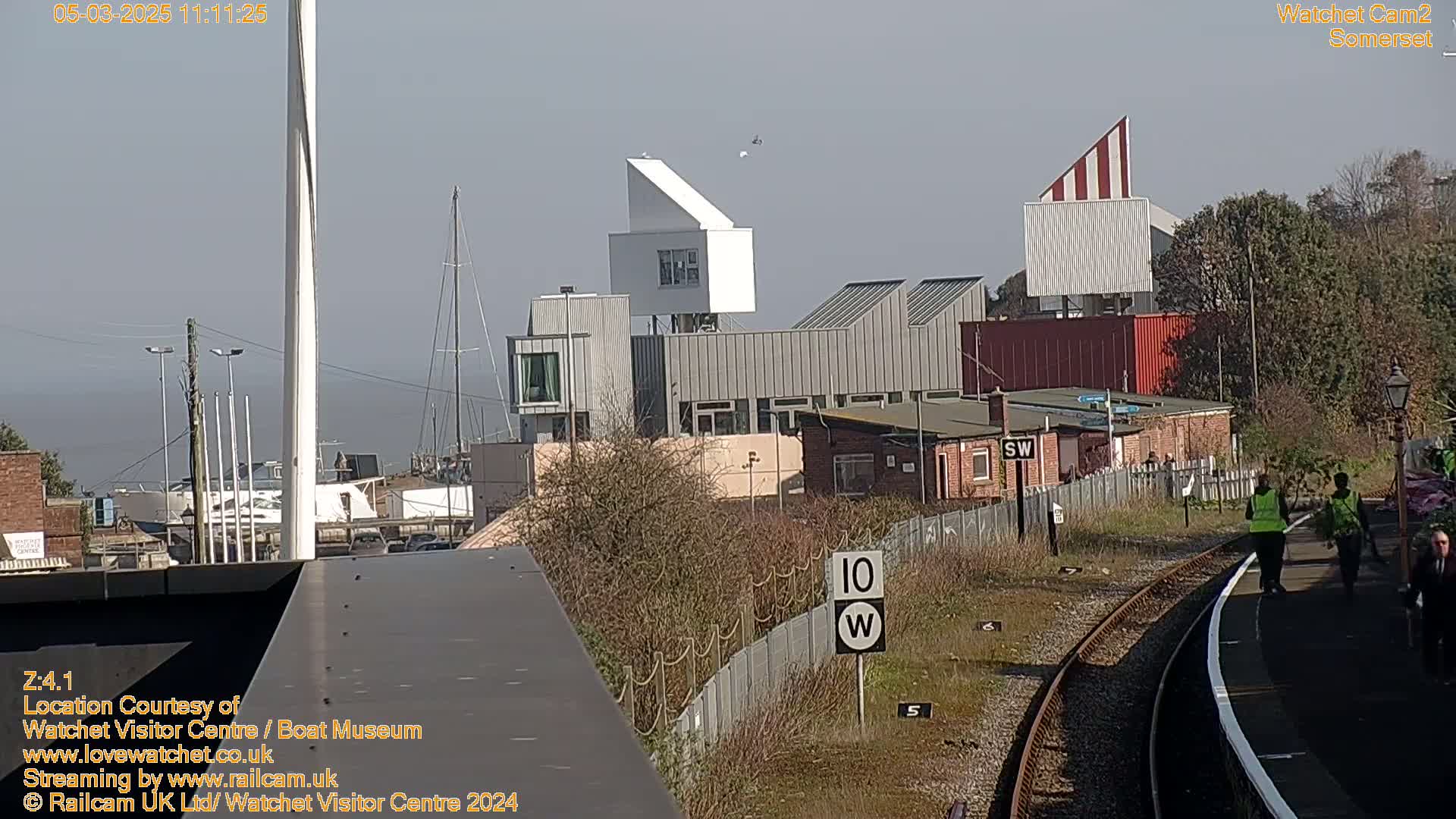 A sunny day reveals a harbor scene with modern buildings, a train track, and several people walking along a platform.