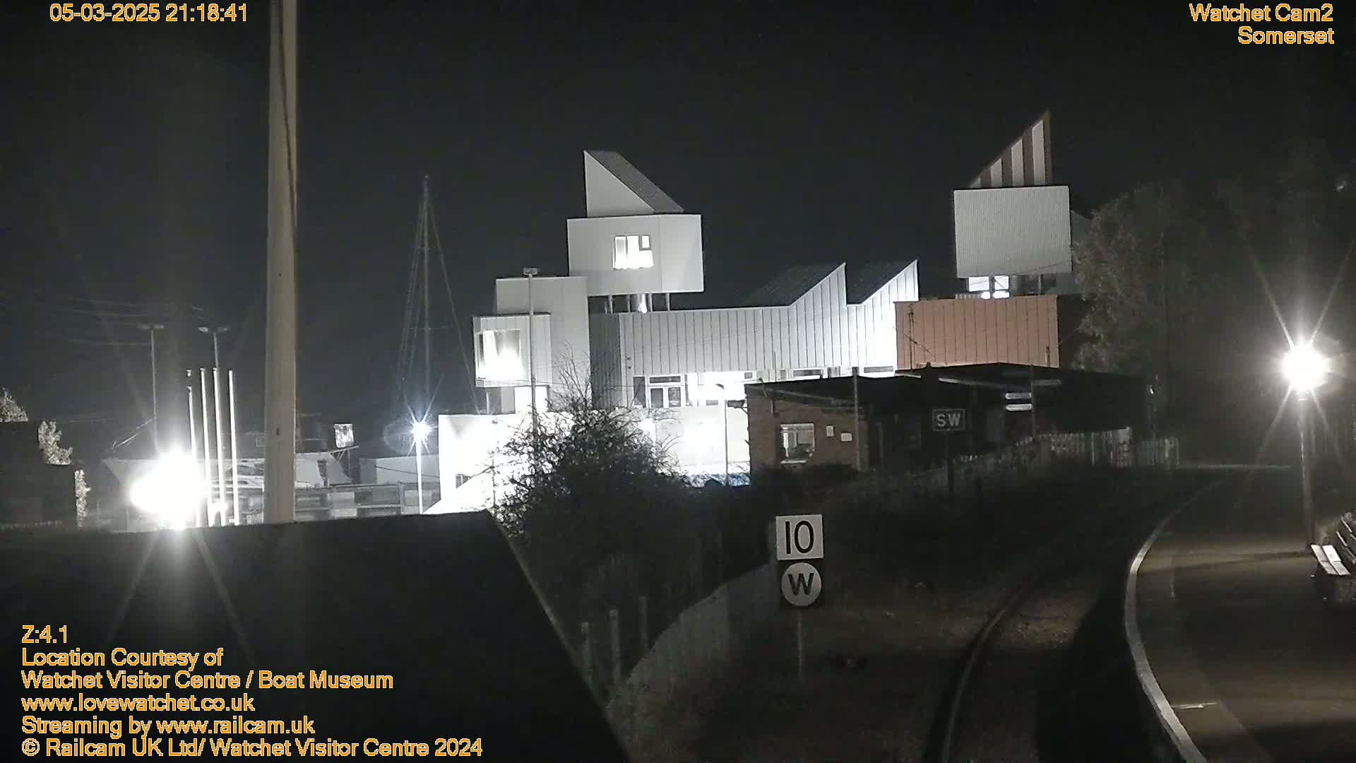 A modern, multi-level white building is illuminated at night next to railroad tracks and some smaller buildings.