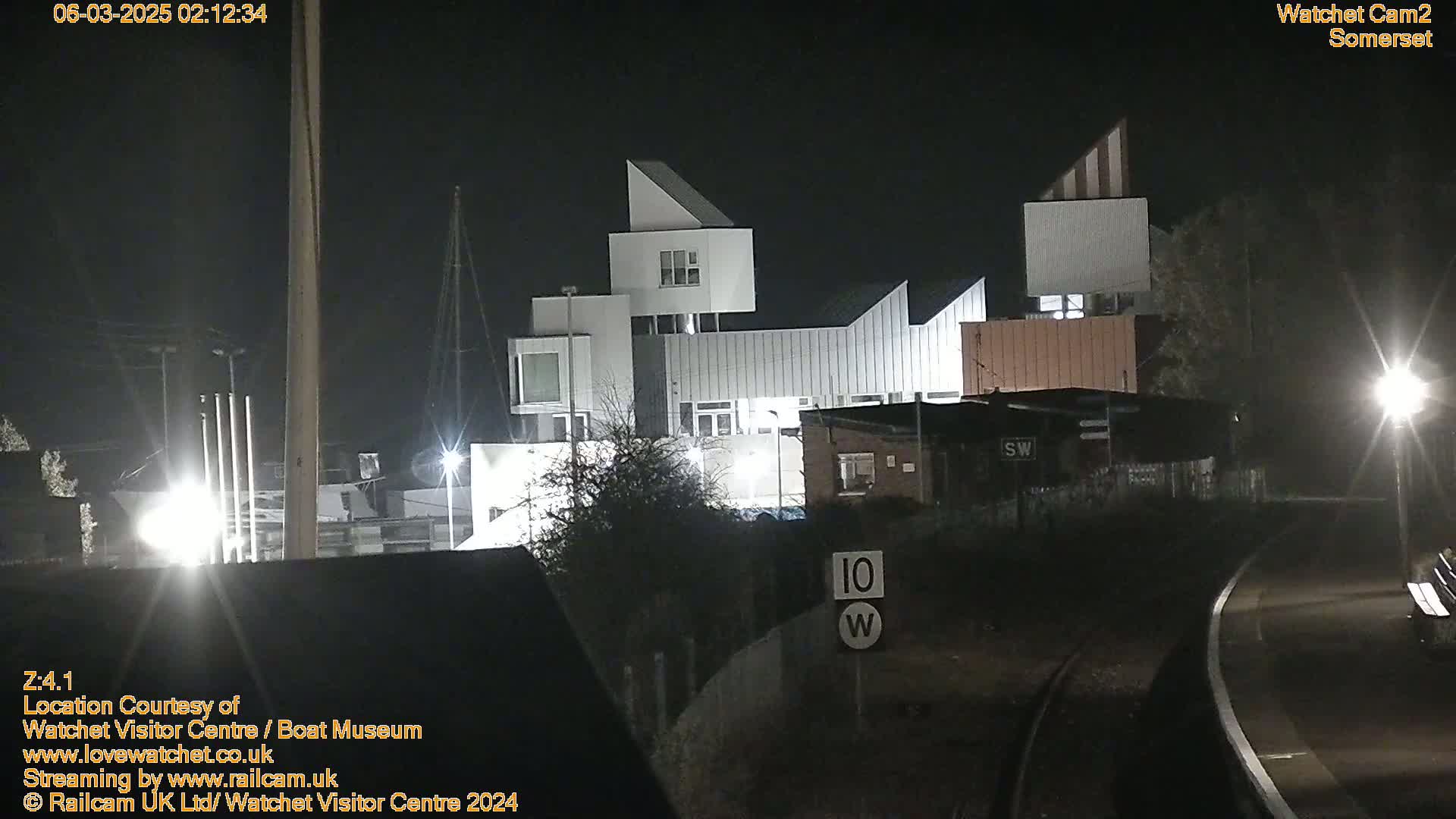 At night, a modern, angular white building complex is illuminated, with a train track and a speed limit sign visible in the foreground.