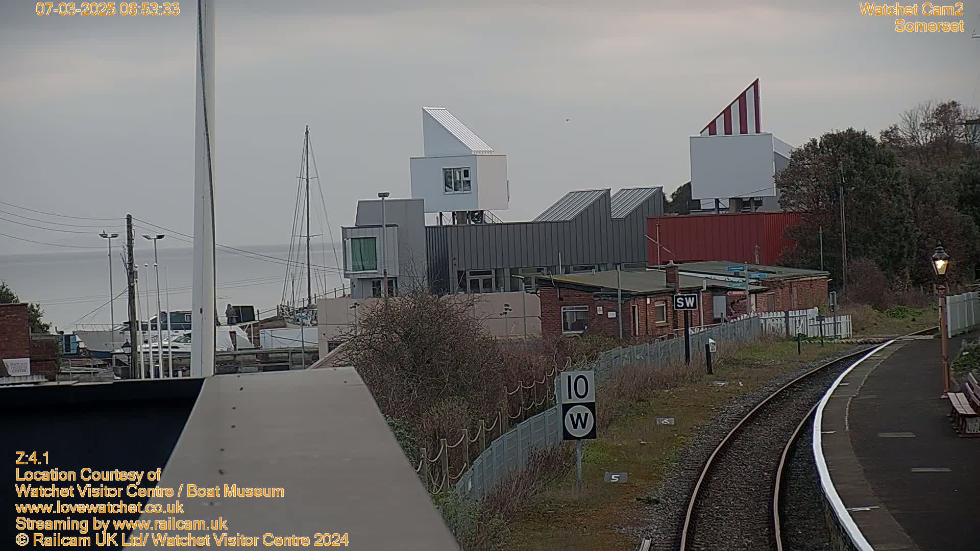 A view of a harbor with modern buildings, a train track, and calm water under an overcast sky.